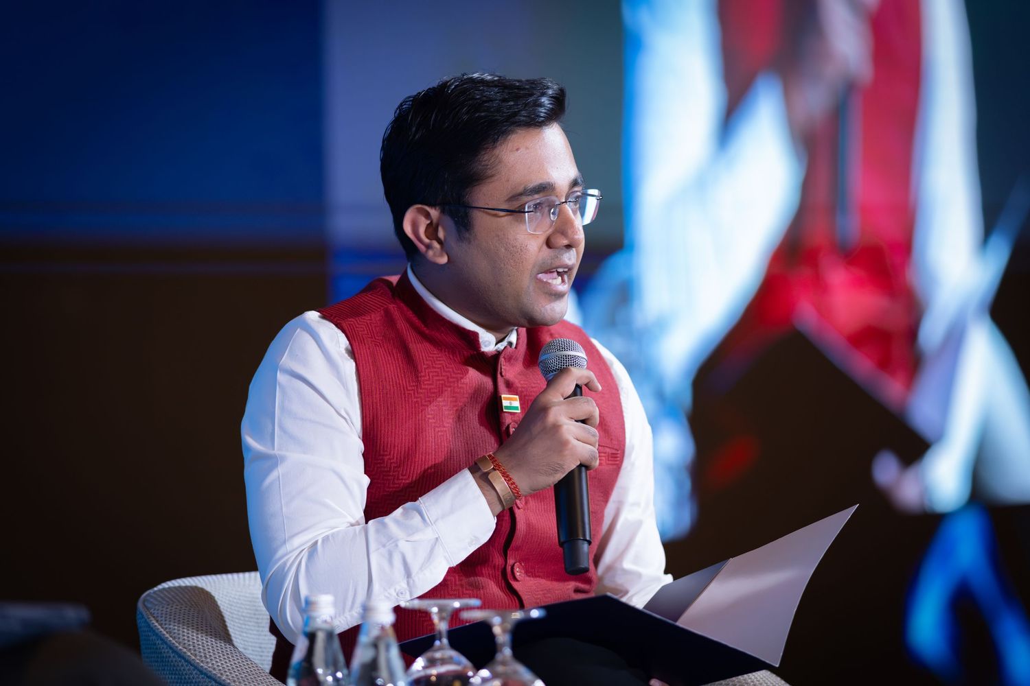 Young man in red vest speaking into microphone at formal conference or panel discussion event.