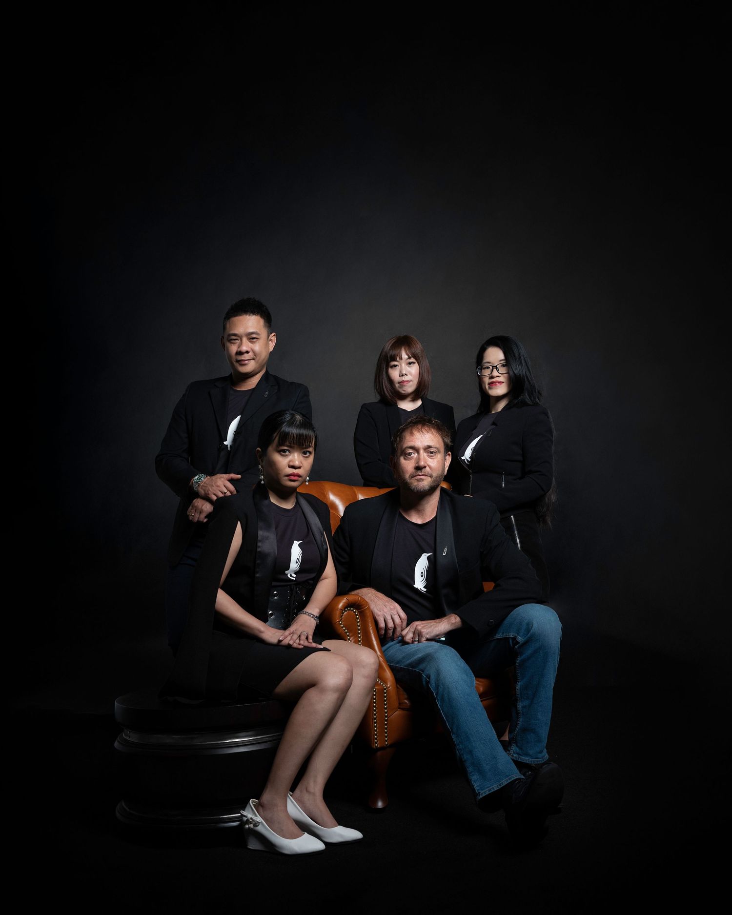 Five people in black outfits pose for a dramatic studio portrait with dark background lighting.