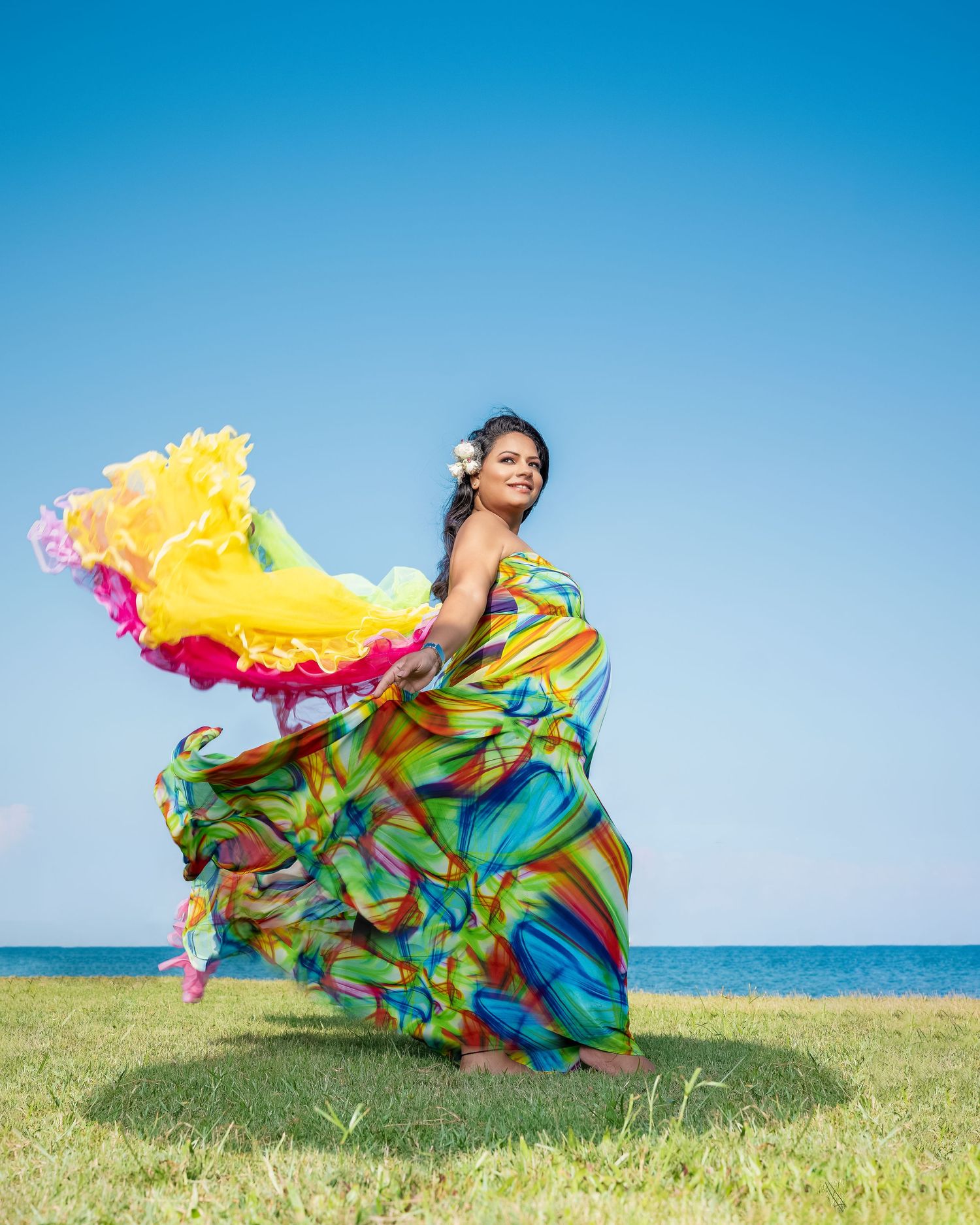 Woman in vibrant multicolored flowing dress dancing outdoors by the ocean under a clear blue sky.