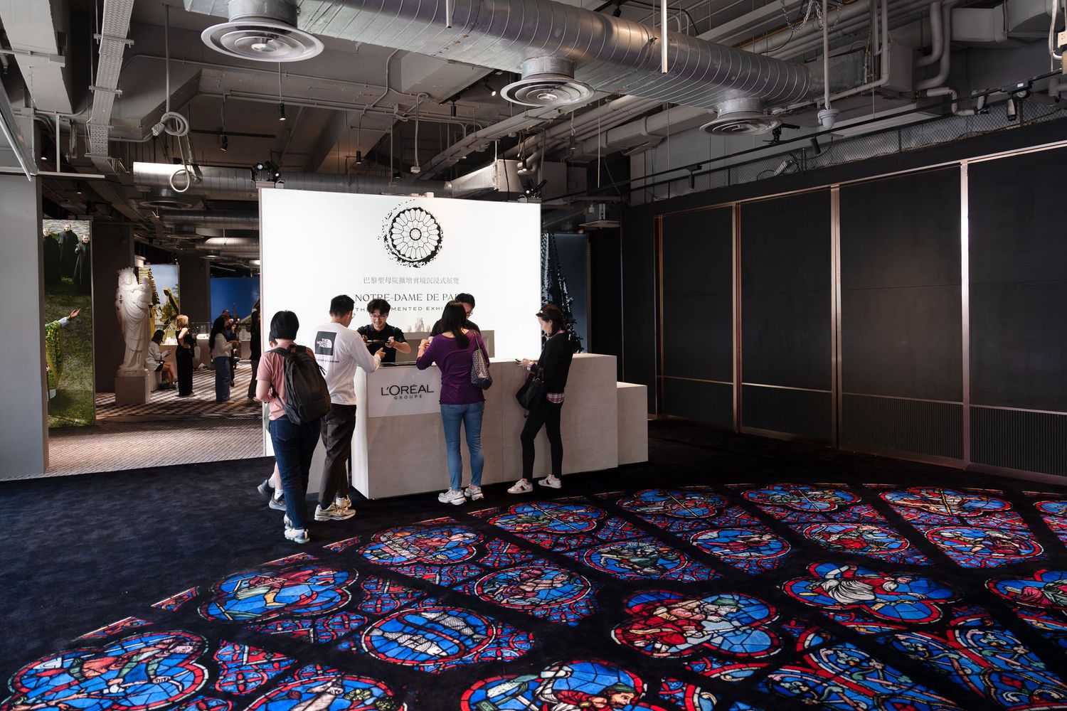 Group of visitors gathered at a reception desk inside a modern exhibition space with colorful stained glass carpet.