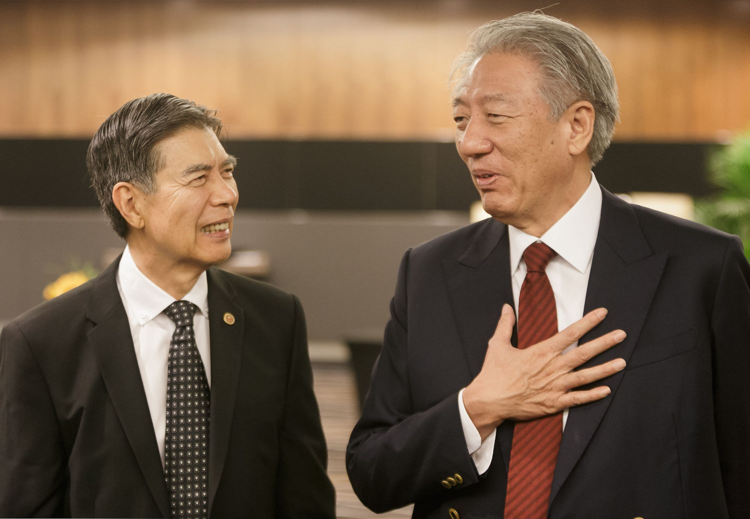 Two Asian businessmen in suits having a conversation, one gesturing with hand on chest in a formal setting.