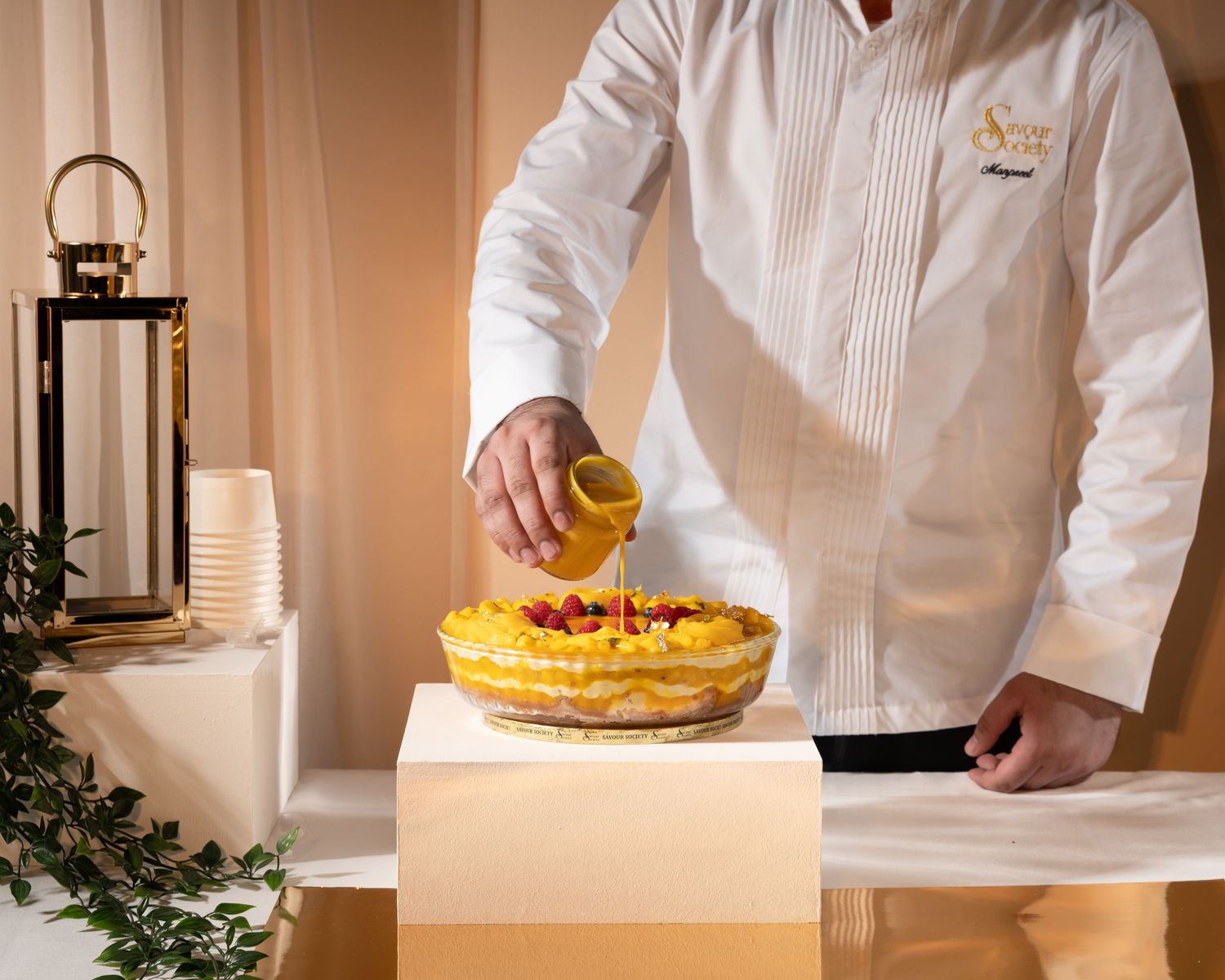 Chef pouring yellow sauce over a layered fruit tart on a white pedestal at an elegant event.