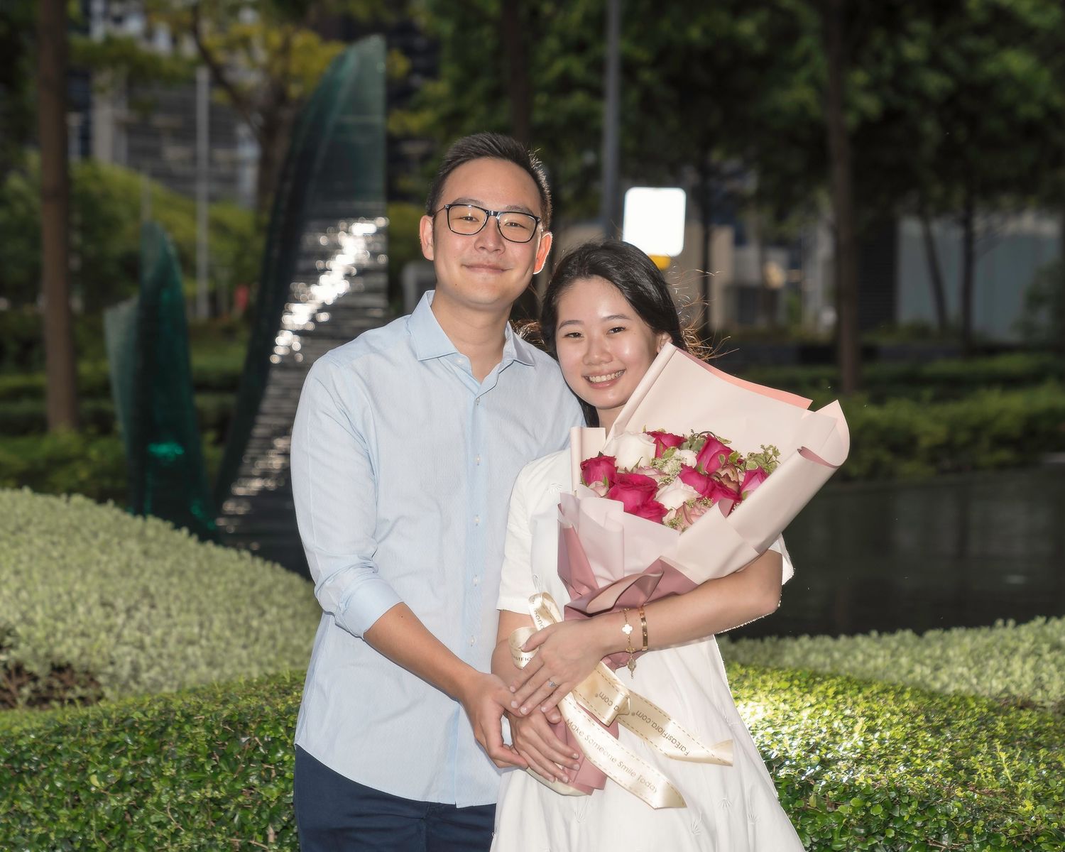 Couple posing outdoors, woman holding pink and red flower bouquet, man in blue shirt, both smiling warmly.