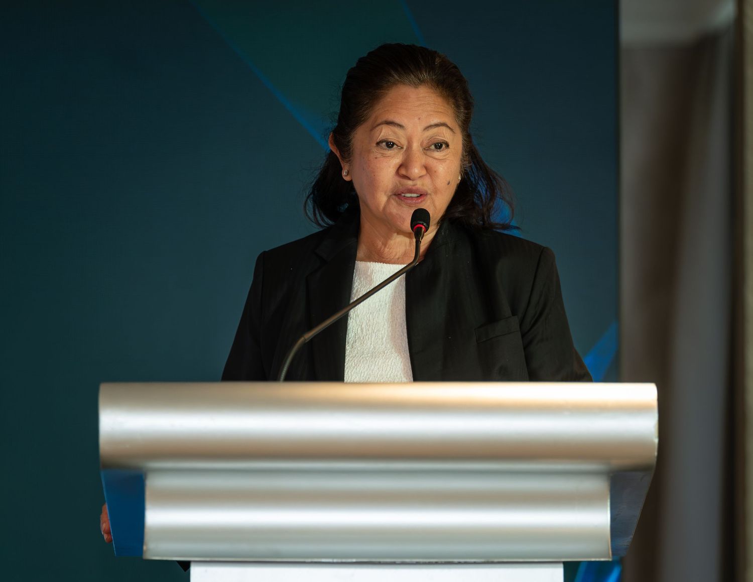 Woman in black blazer speaking at a silver podium during a professional conference or corporate event.