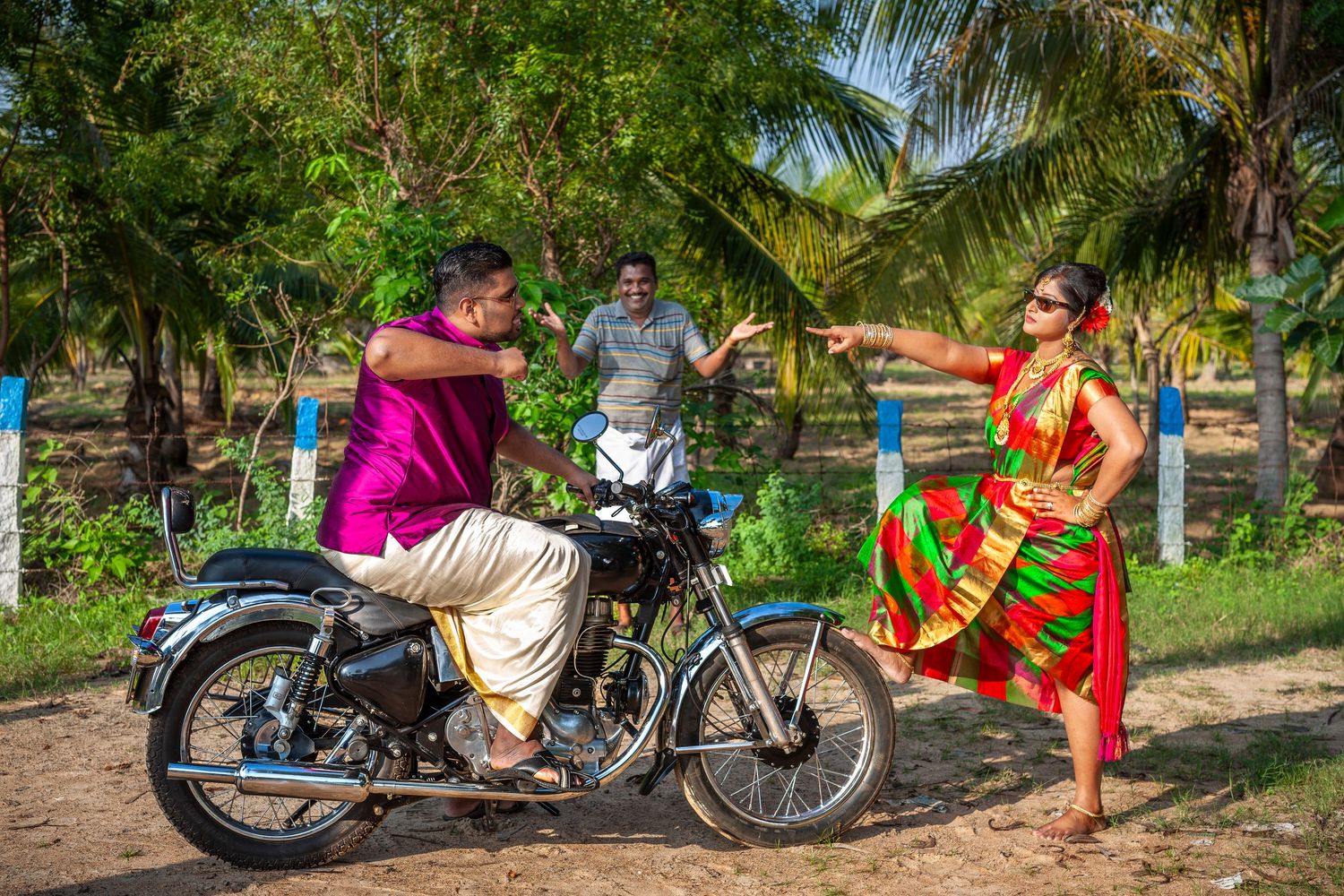Man on Royal Enfield motorcycle with two people in colorful traditional Indian attire near palm trees.
