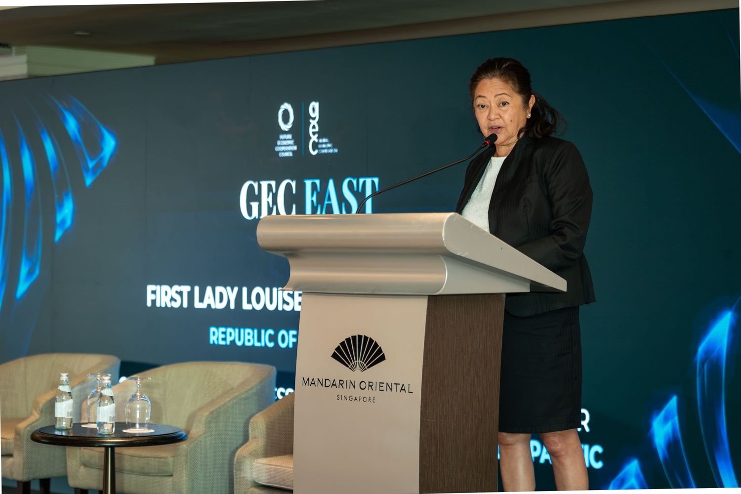 First Lady Louise speaks at GEC East podium at Mandarin Oriental, representing the Republic in a professional black suit.
