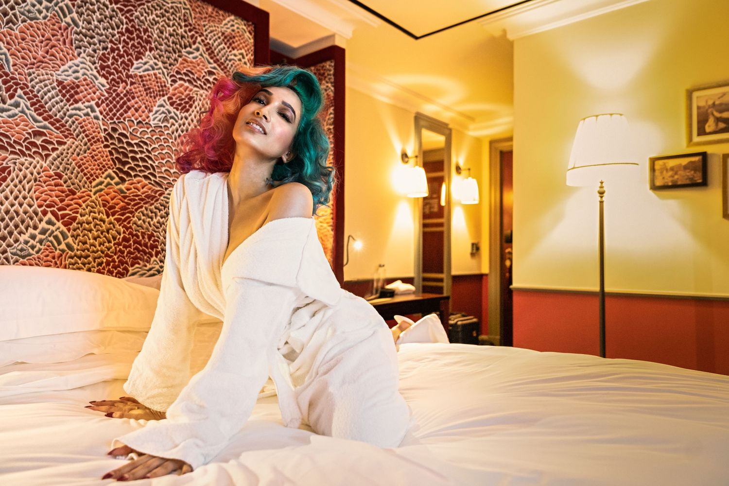 Woman with colorful hair in white robe sitting on hotel bed, smiling in a warmly lit room.