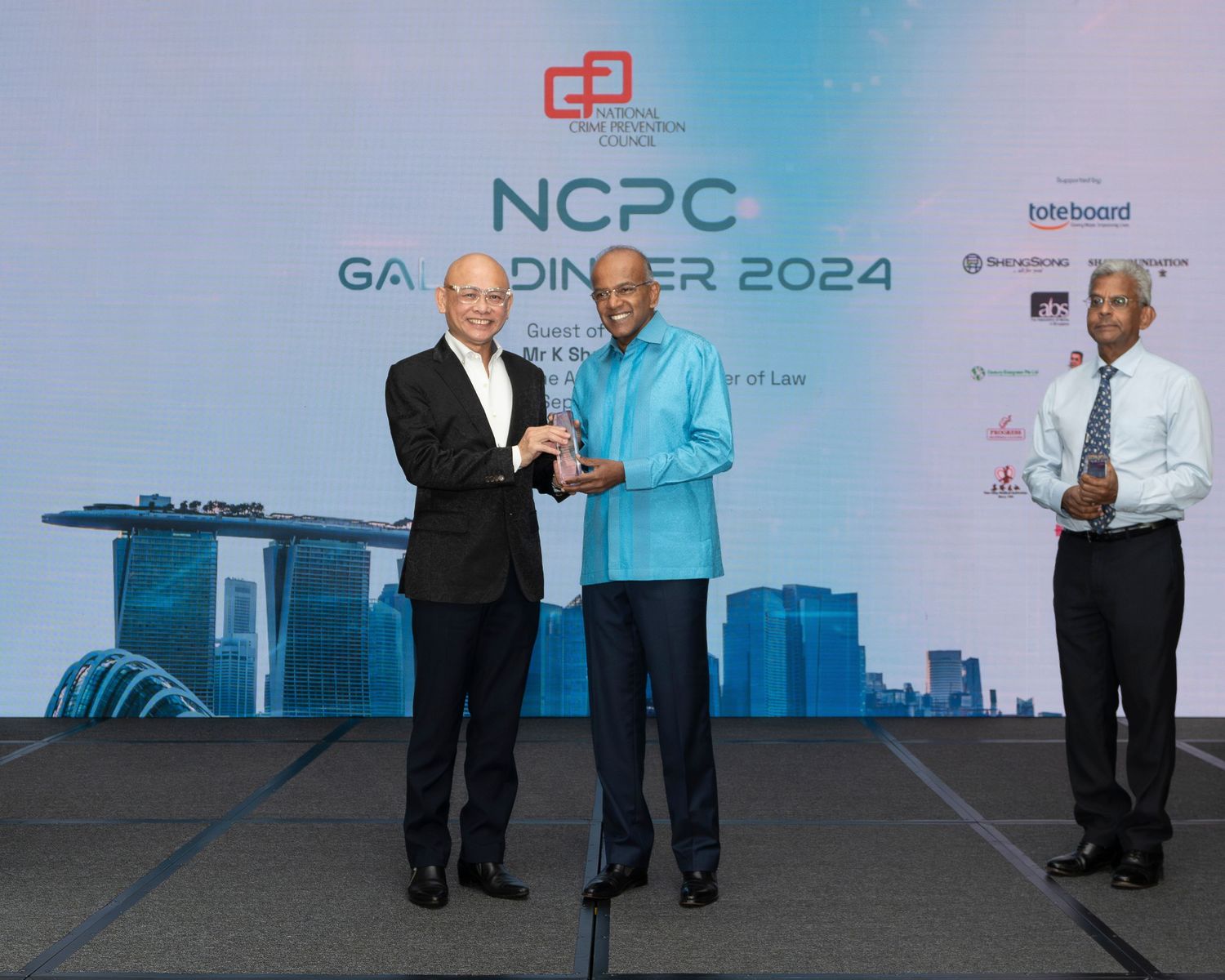 Three men in formal attire exchange award at NCPC Gala Dinner 2024 at Marina Bay Sands Singapore.