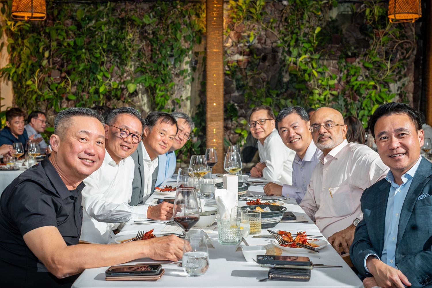 Group of men smiling and dining together at an upscale restaurant with lush green wall backdrop.