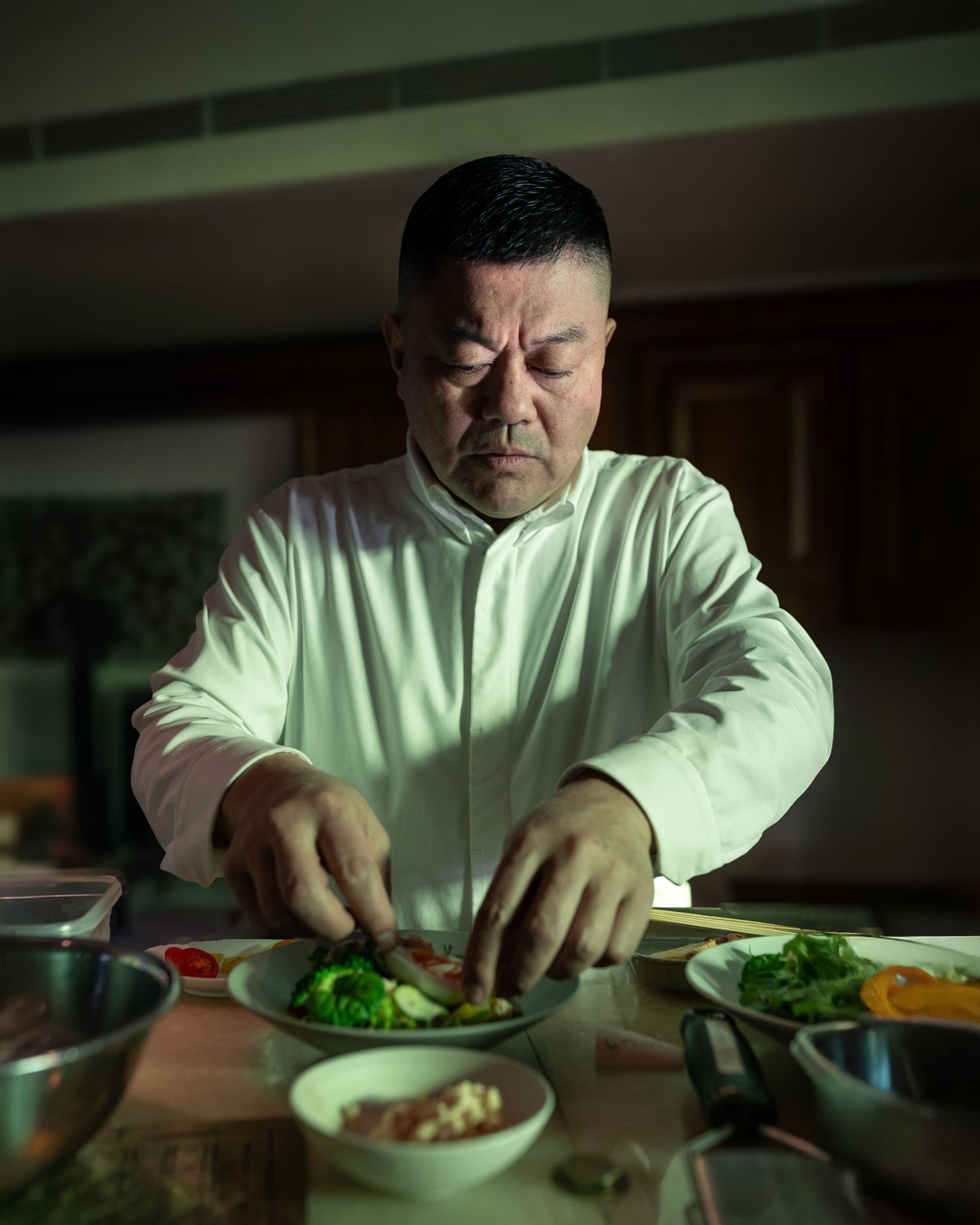 A chef in a white uniform carefully plates a dish with broccoli and meat on a dark kitchen counter.
