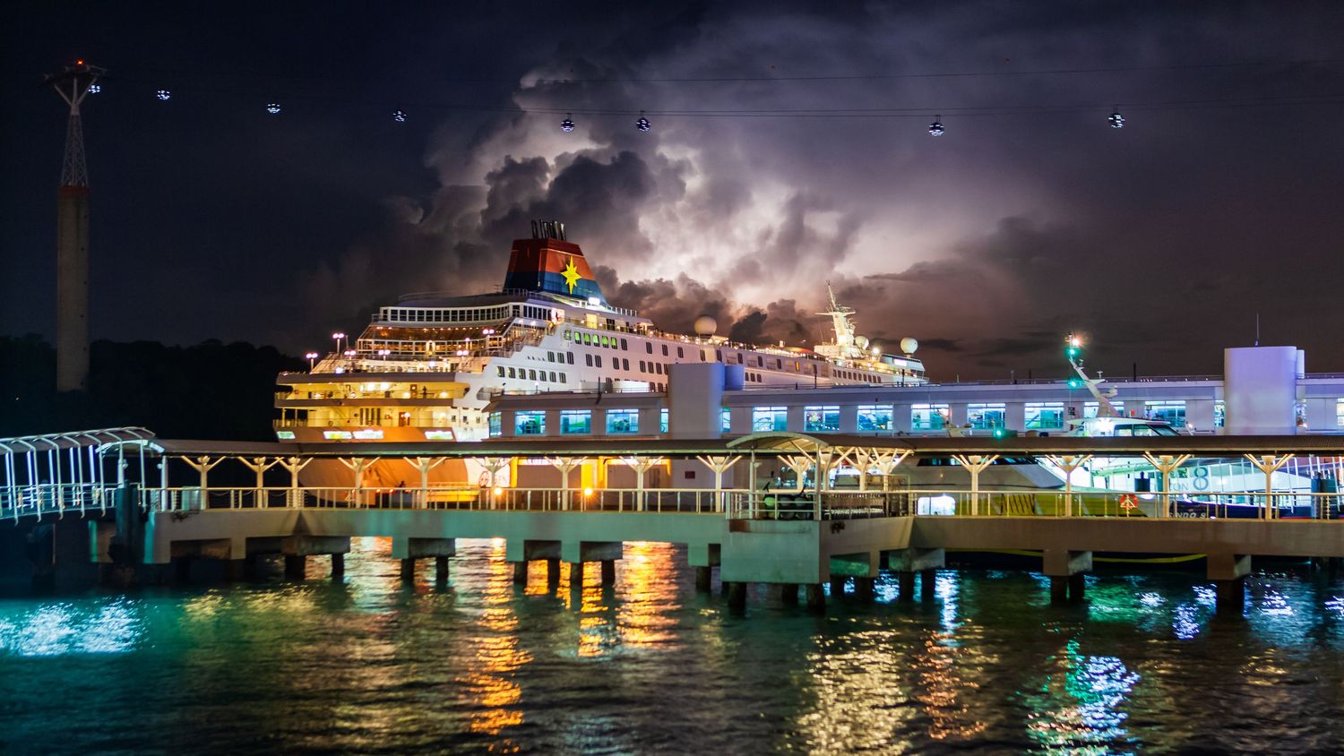 A large cruise ship docked at a illuminated pier at night with dramatic lightning storm in the background.