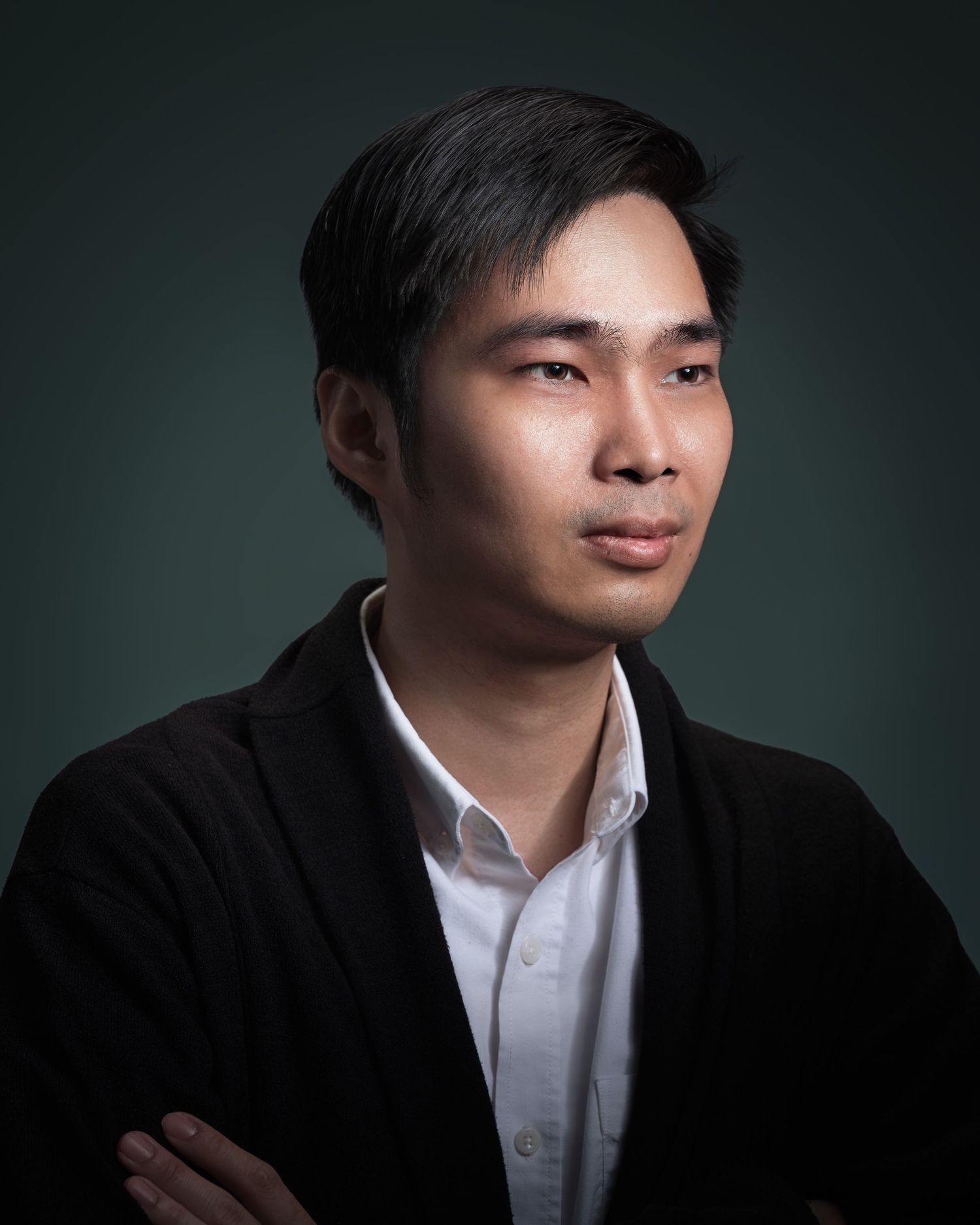 Young Asian man in black cardigan with arms crossed in professional studio portrait against dark green background.