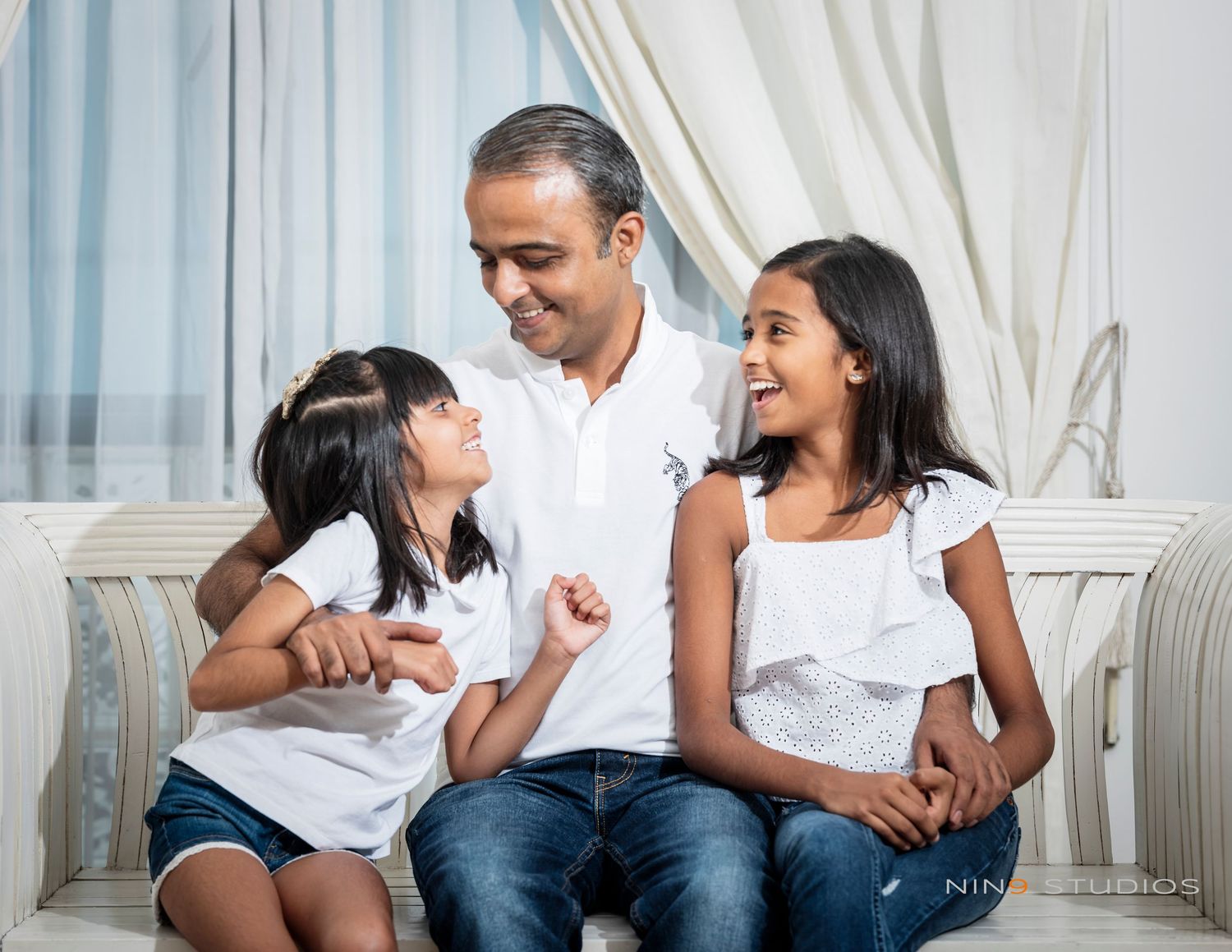 A man smiles warmly with two young girls in white tops, sharing a joyful moment on a couch.