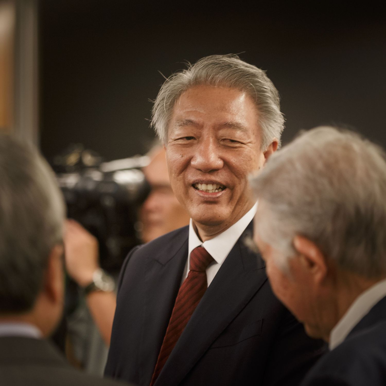 Smiling senior Asian businessman in dark suit at formal event, engaging with colleagues in professional setting.