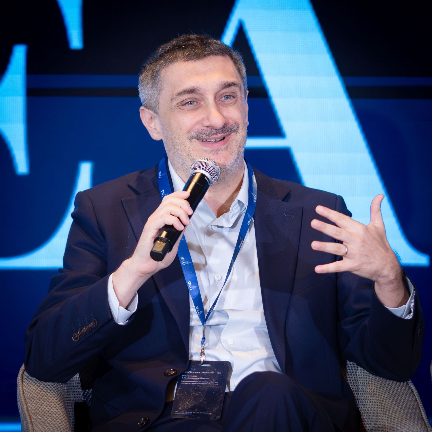 Man in suit speaking at conference panel holding microphone with blue background.