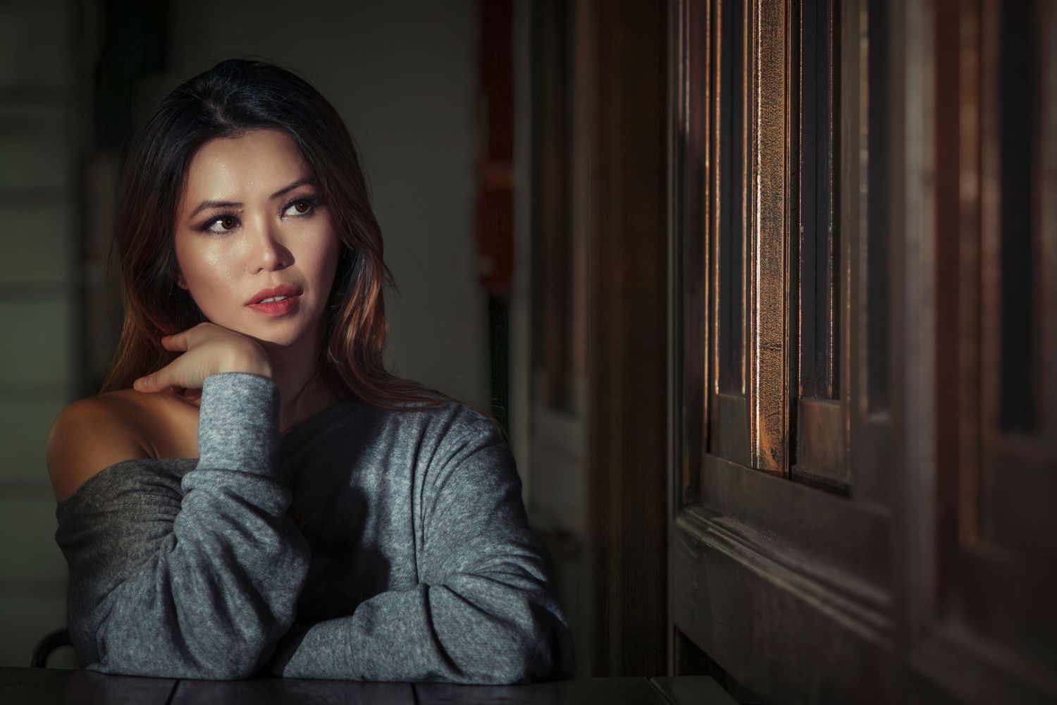 Young Asian woman in gray off-shoulder sweater gazes thoughtfully beside a wooden door in moody lighting.