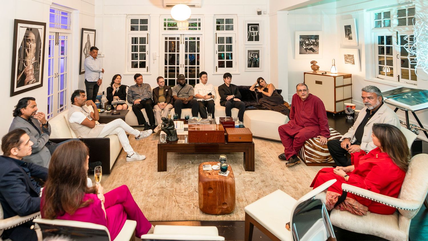 A diverse group gathers in an elegant white living room with a Buddhist monk in red robes during an intimate discussion.