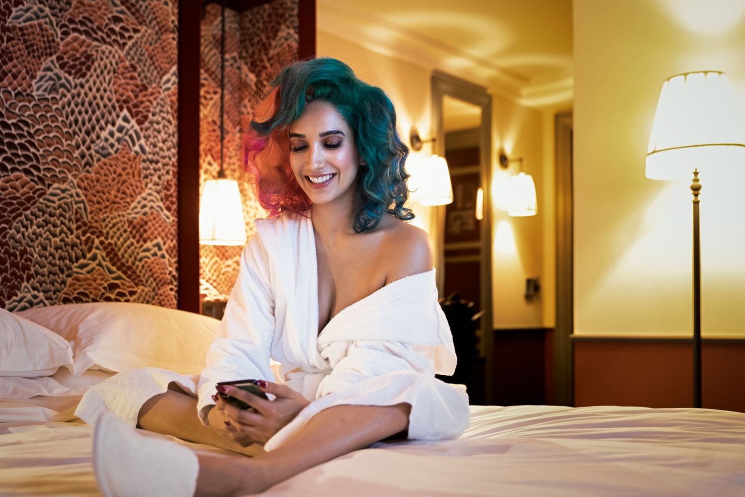 Woman with teal curly hair in white robe sitting on hotel bed smiling at smartphone.