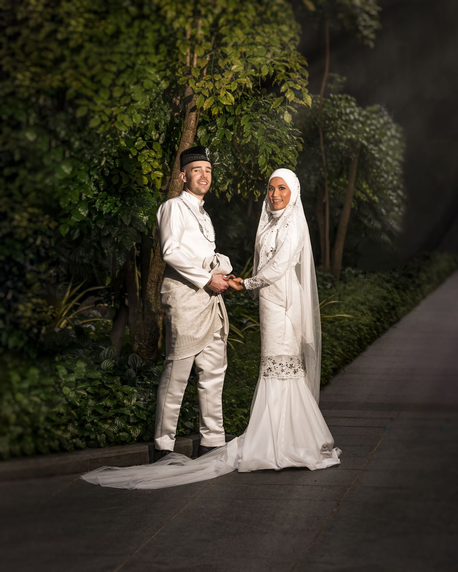Muslim couple in white traditional attire holding hands outdoors, with lush green foliage backdrop, wedding portrait.