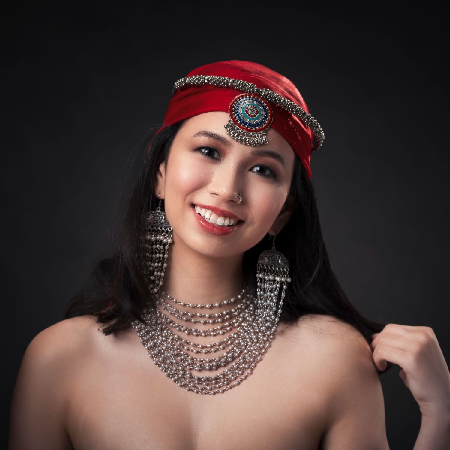 Smiling woman wearing red headpiece and silver tribal jewelry poses against a dark studio background.