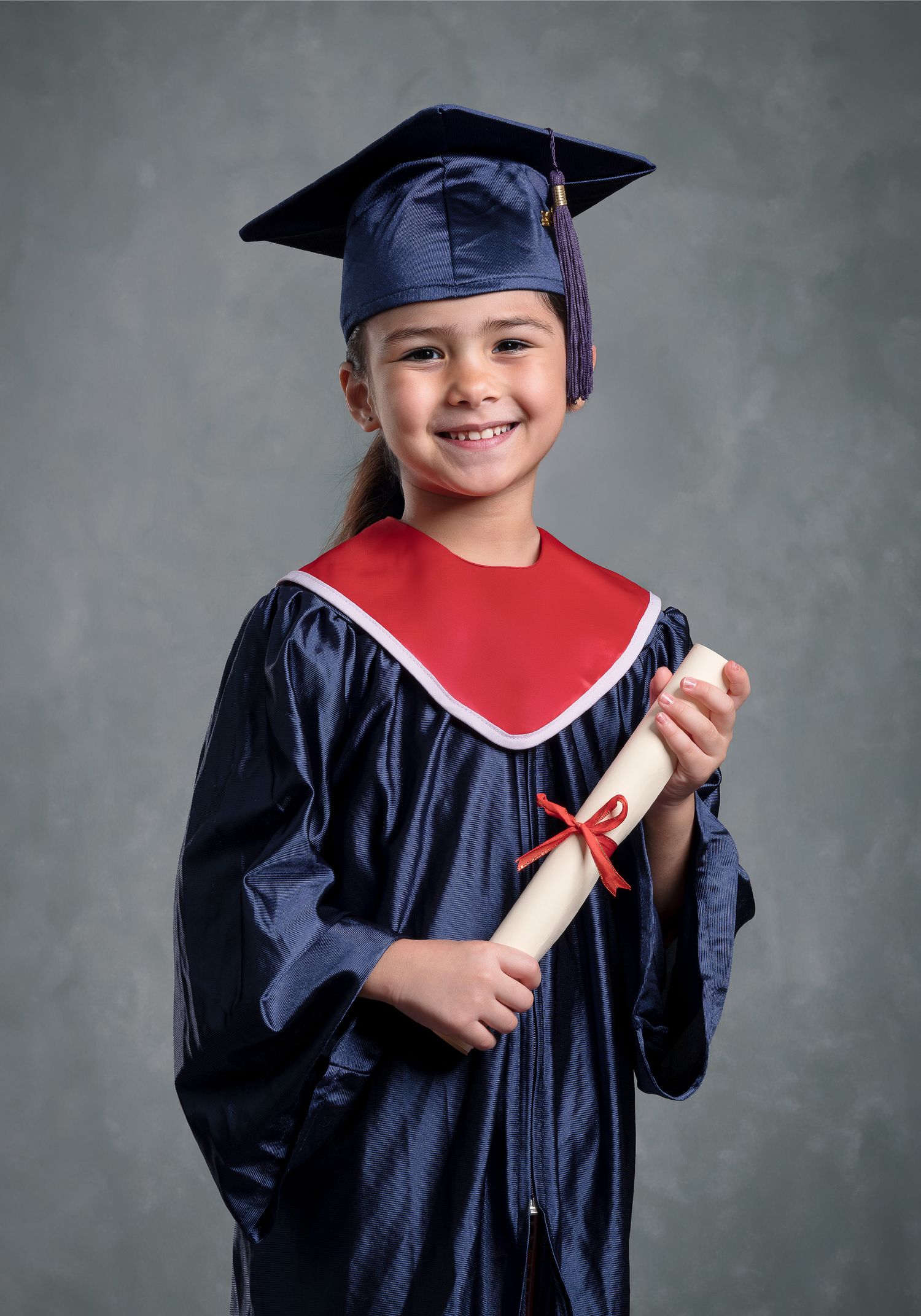 Smiling young girl in navy blue graduation cap and gown holding a diploma scroll with red ribbon.