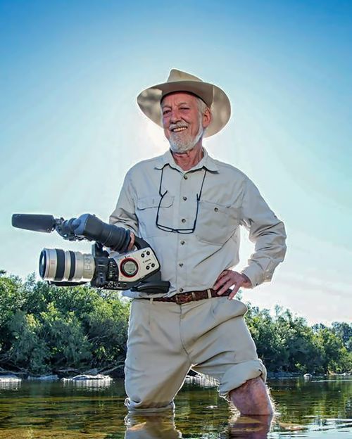 Smiling man in khaki outfit and cowboy hat holding professional video camera while standing in shallow river water.