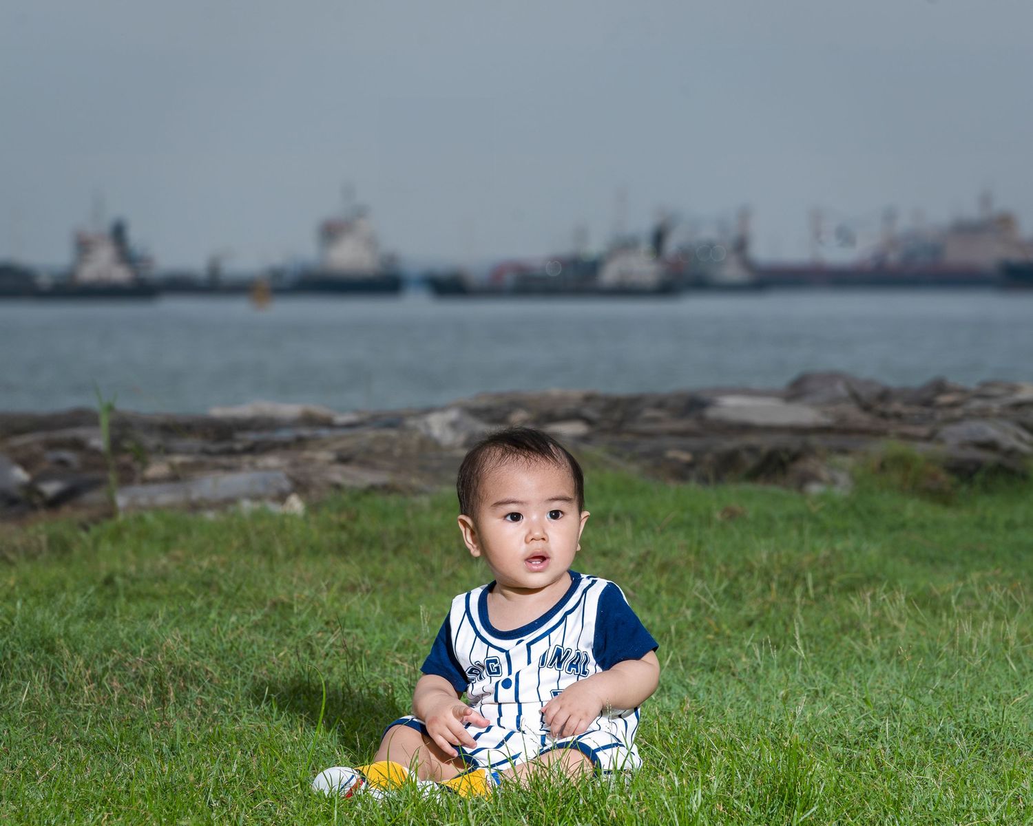 A baby boy in a navy romper sits on green grass by the waterfront with large ships visible in the background.