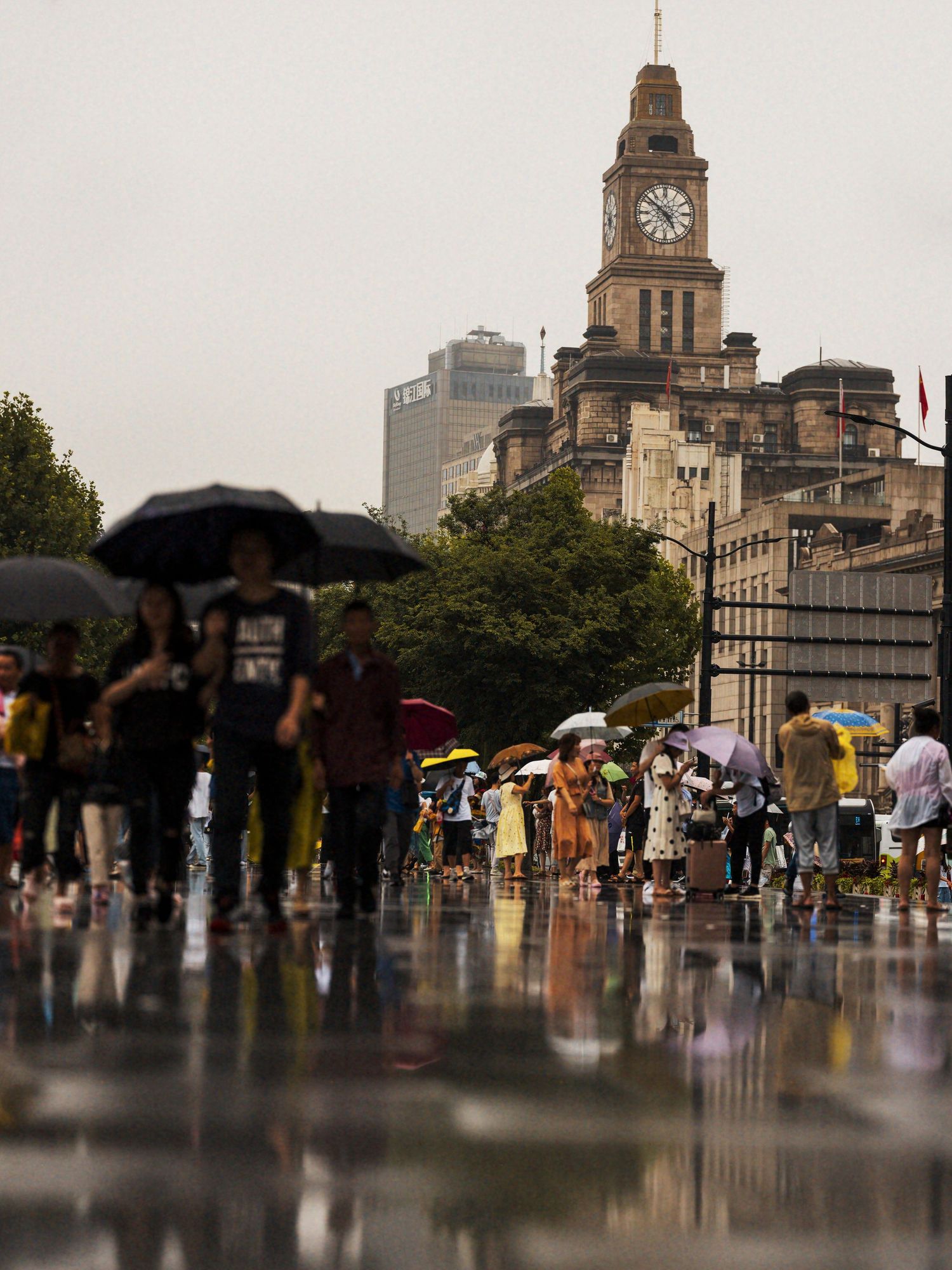 Crowds with colorful umbrellas walk along the Bund in Shanghai on a rainy day, with a historic clock tower in the background.
