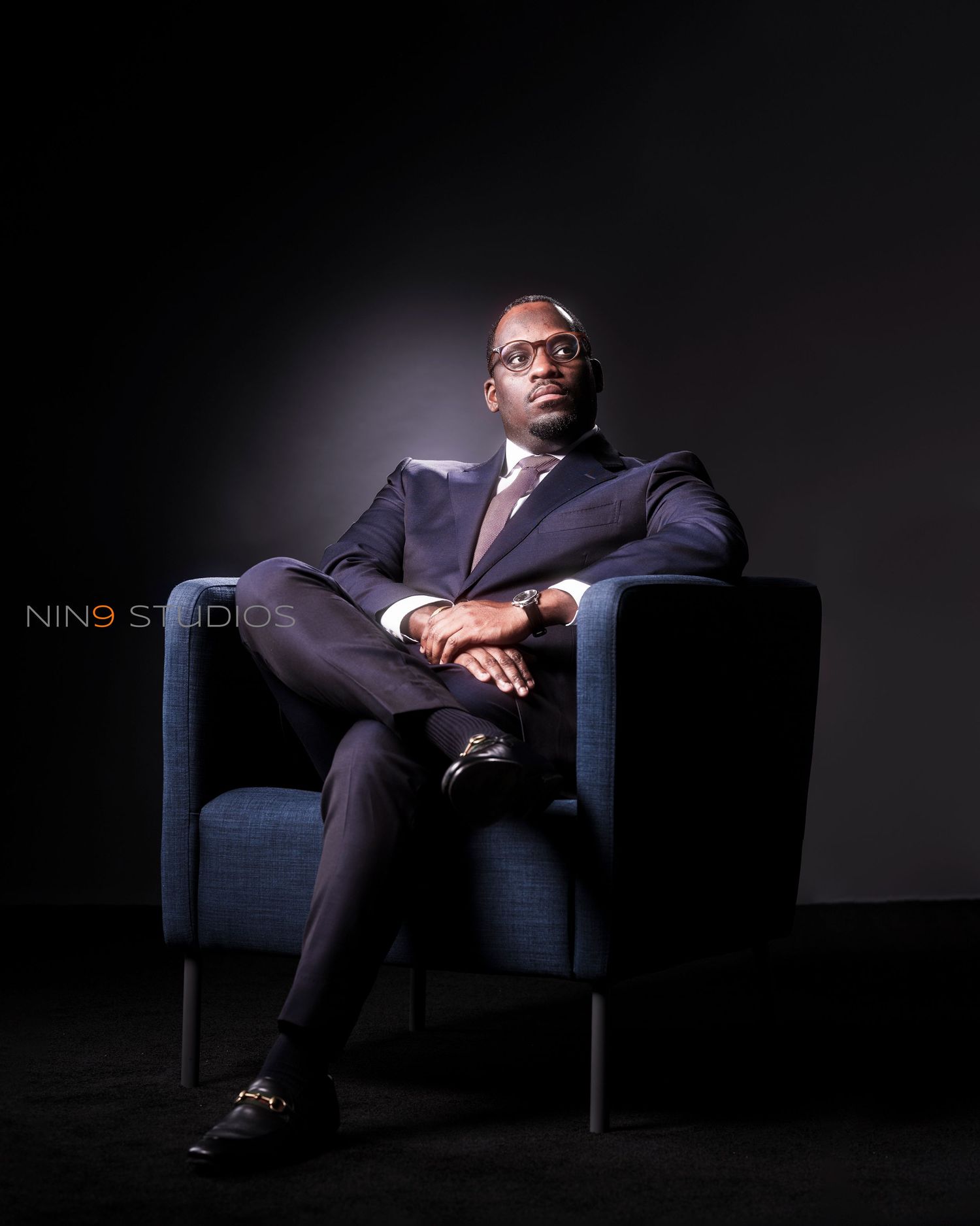 A confident man in a navy suit sits cross-legged in a blue chair against a dark background.