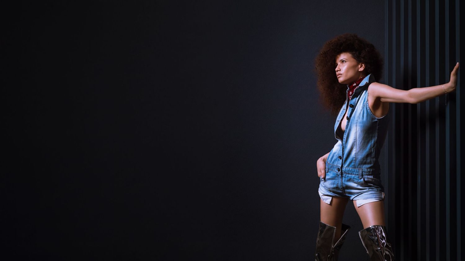 A stylish woman with an afro poses confidently in denim shorts and boots against a dark background.
