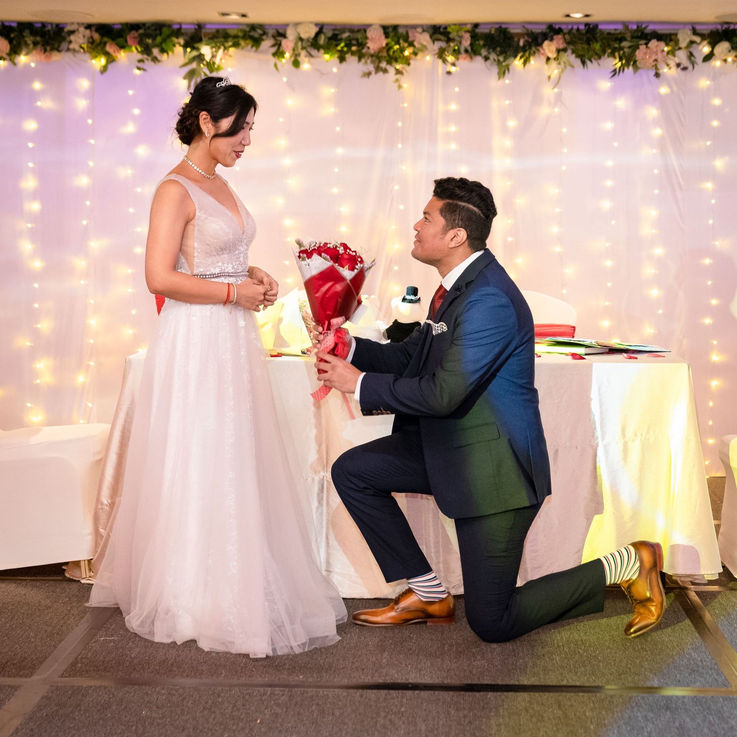 A groom kneels presenting red roses to his bride in a white gown before a glowing fairy light backdrop.