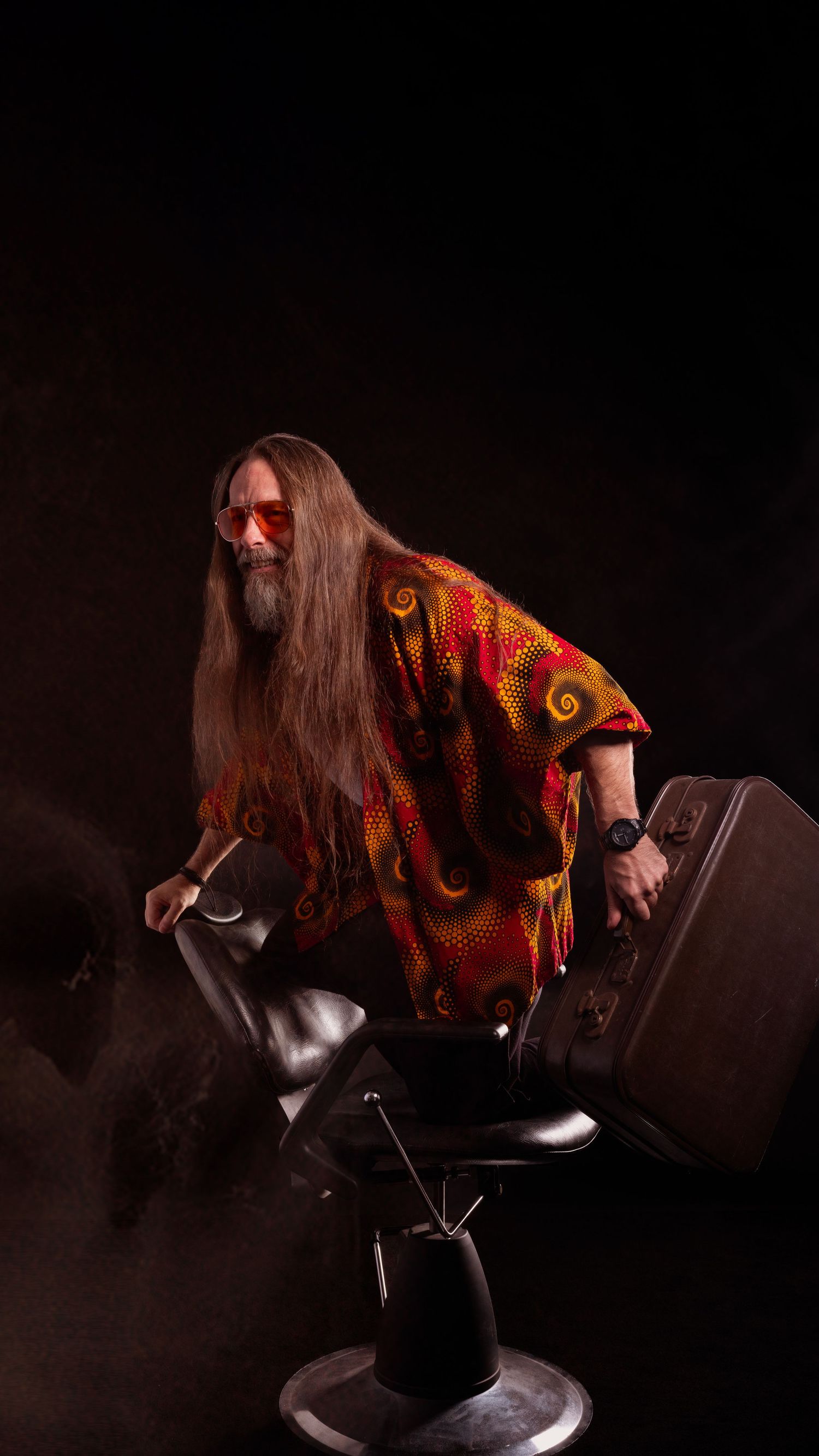 Long-haired man in colorful shirt and red sunglasses sitting in a barber chair against dark background.