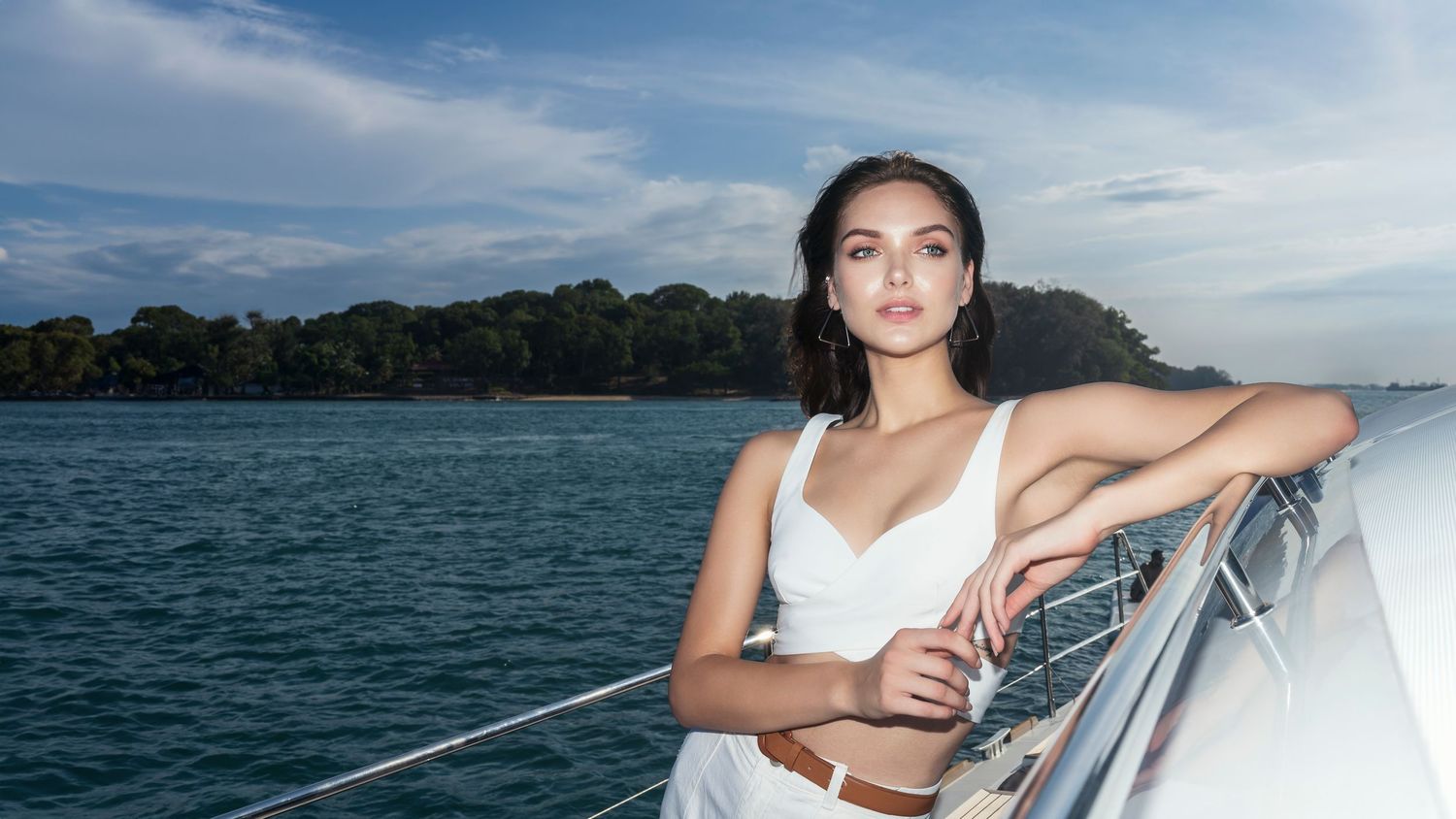 Young woman in white bikini top posing on luxury yacht railing with tropical coastline and blue sky backdrop.