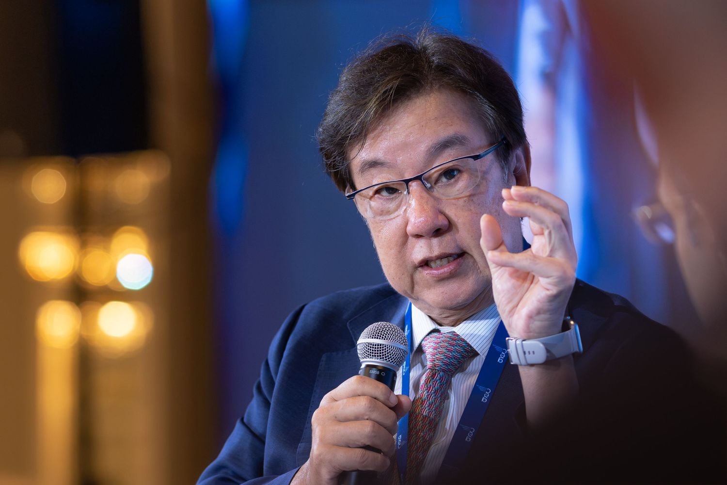 An Asian man in a suit gestures while speaking into a microphone at a formal conference panel.