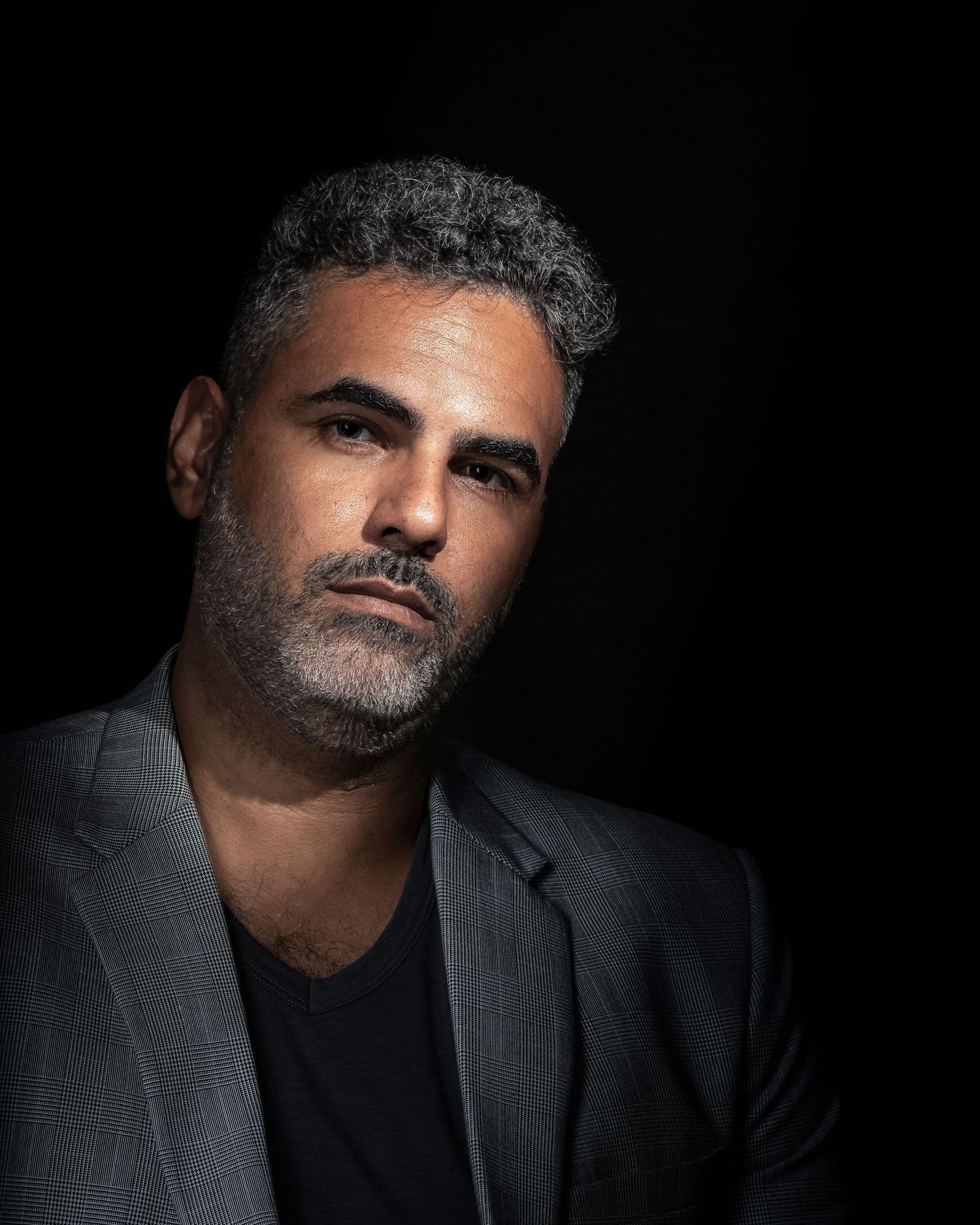 Middle-aged man with salt-and-pepper hair and beard wearing a gray blazer poses against a dark background.