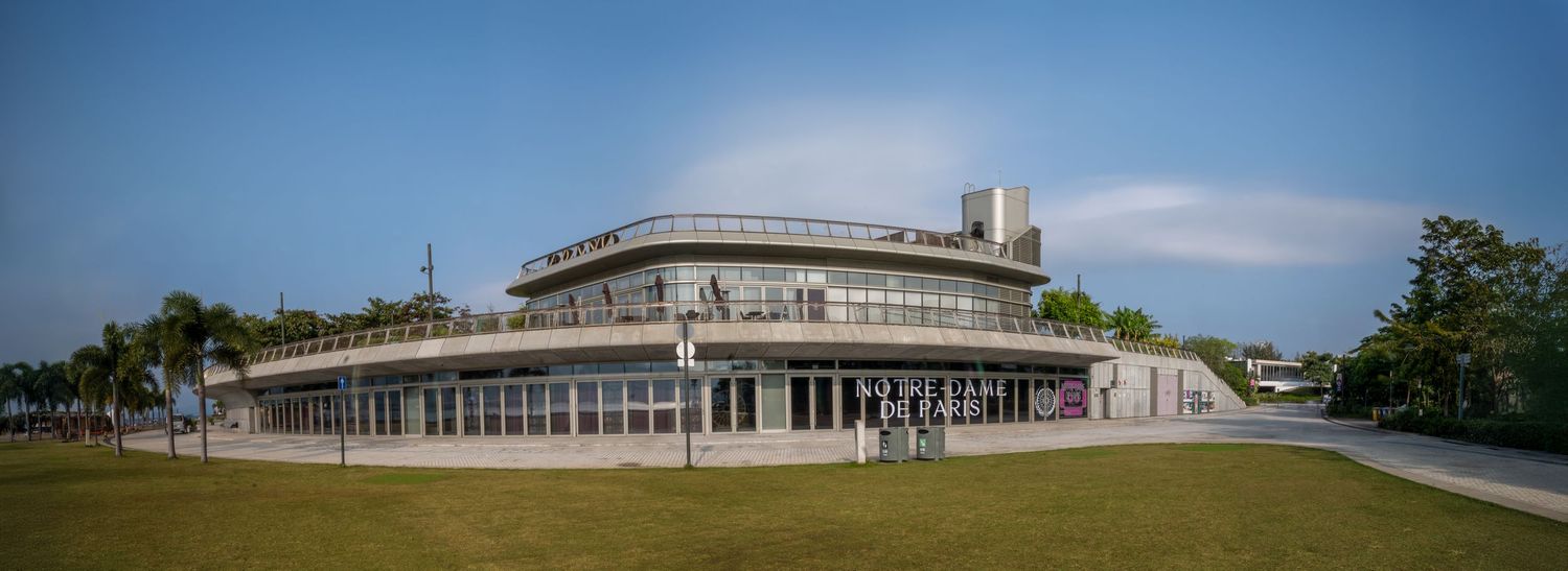 Modern glass and steel building with curved architecture surrounded by lush green trees under a clear blue sky.