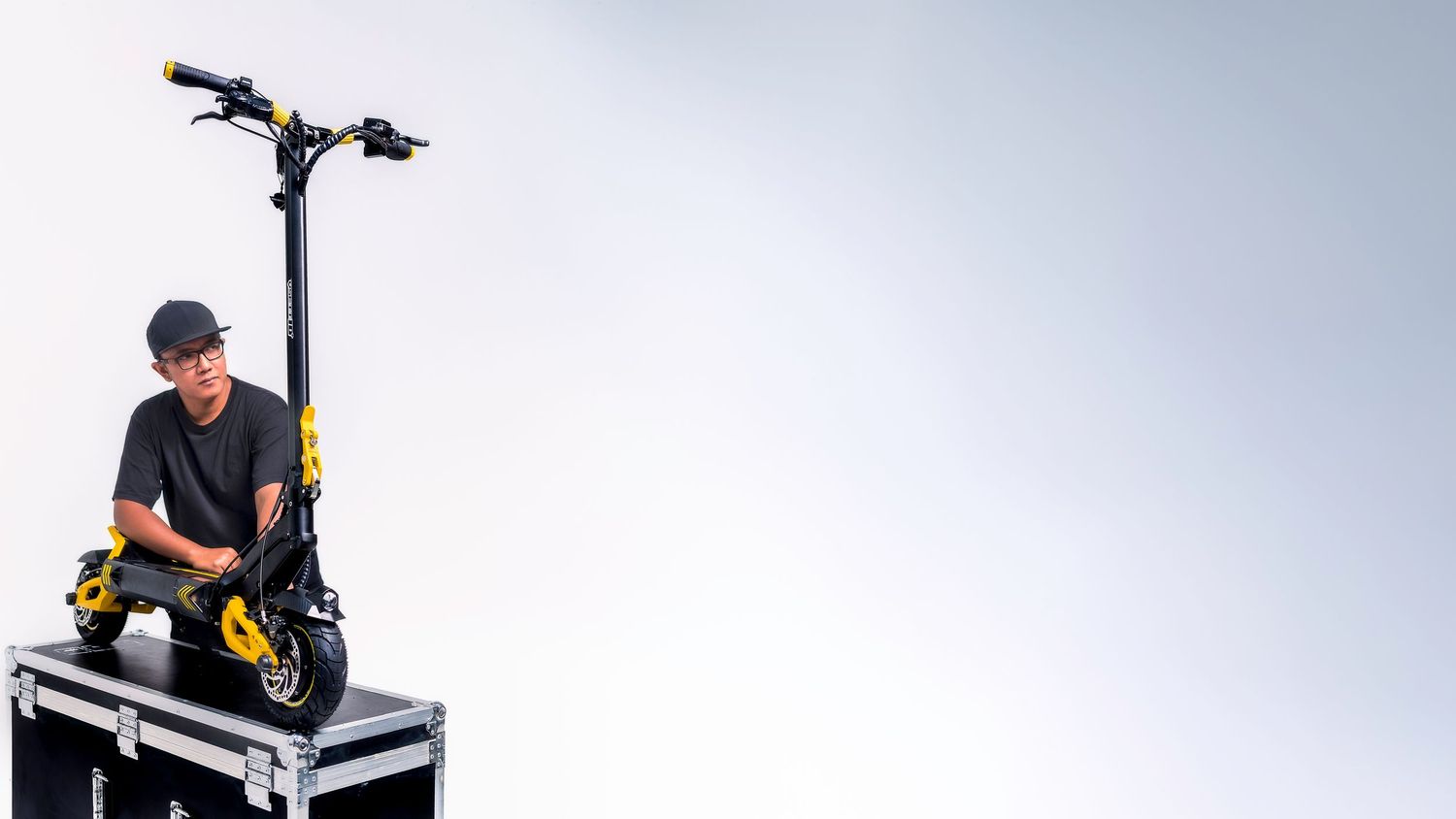Man in black outfit sitting on a yellow and black electric scooter displayed on a flight case against white background.