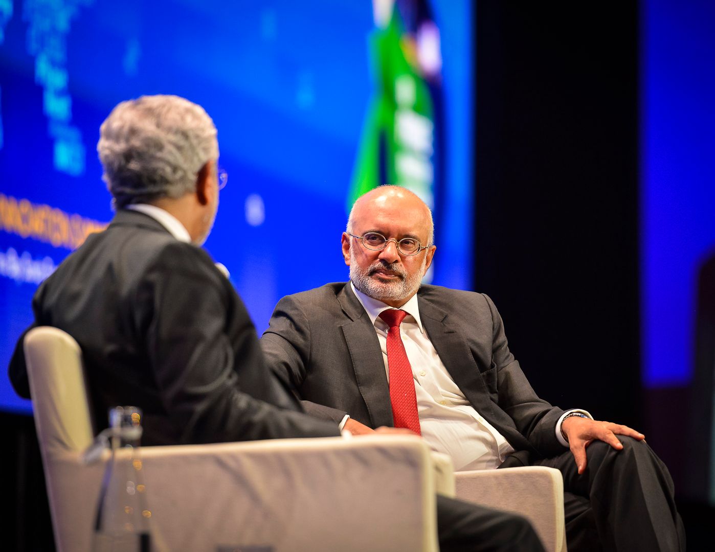 Two suited executives engage in discussion at a formal business conference on stage.