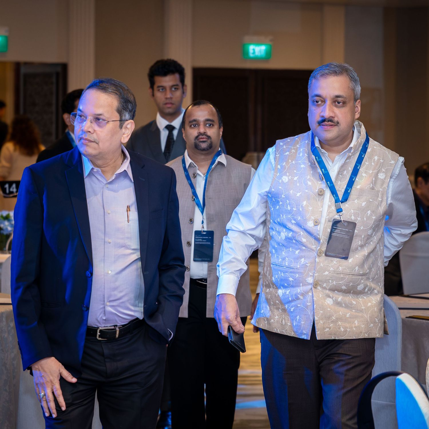 Three men in formal attire walk through a conference venue, two wearing lanyards at a professional event.