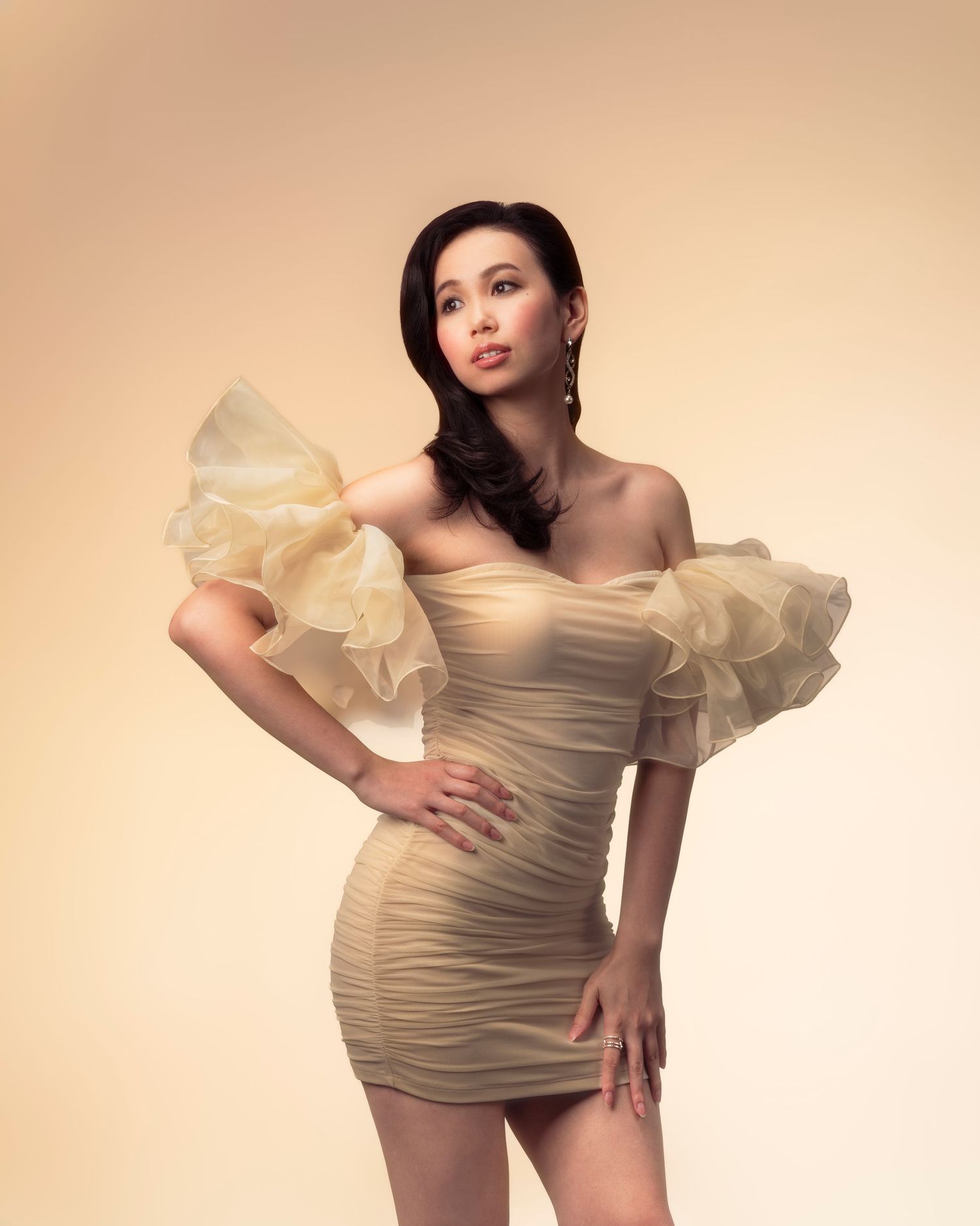 Woman in elegant cream ruched mini dress with dramatic organza ruffle sleeve, posing against a warm beige background.