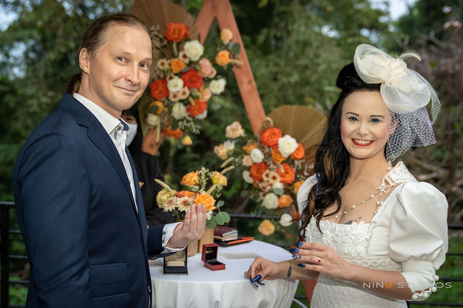 Couple in wedding attire exchanging rings outdoors with orange floral arrangement backdrop.