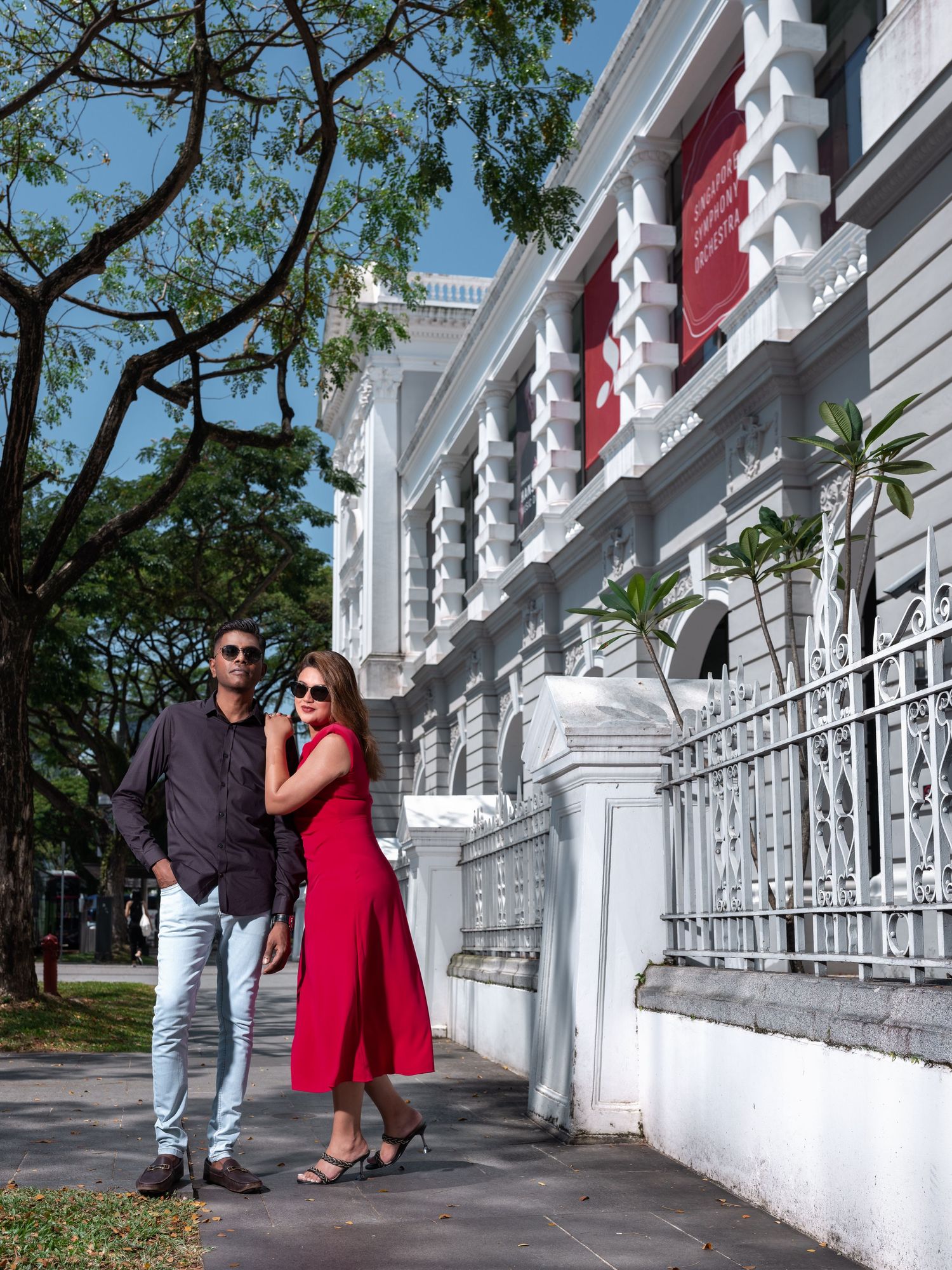 Couple posing in front of a white colonial building, woman in red dress, man in black shirt, sunny day.