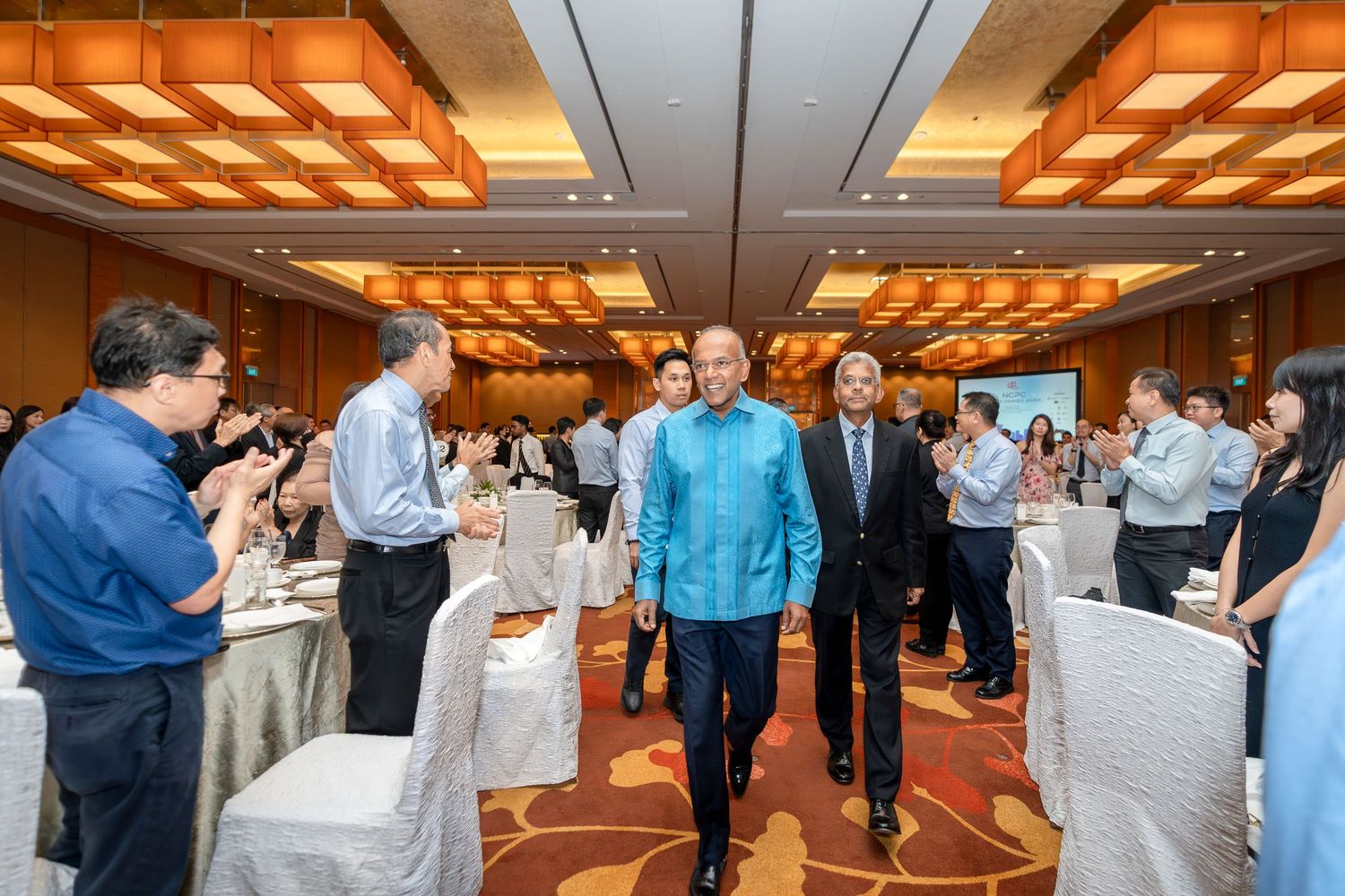 Guests mingle at a formal banquet hall as two men in business attire walk through the elegant event space.