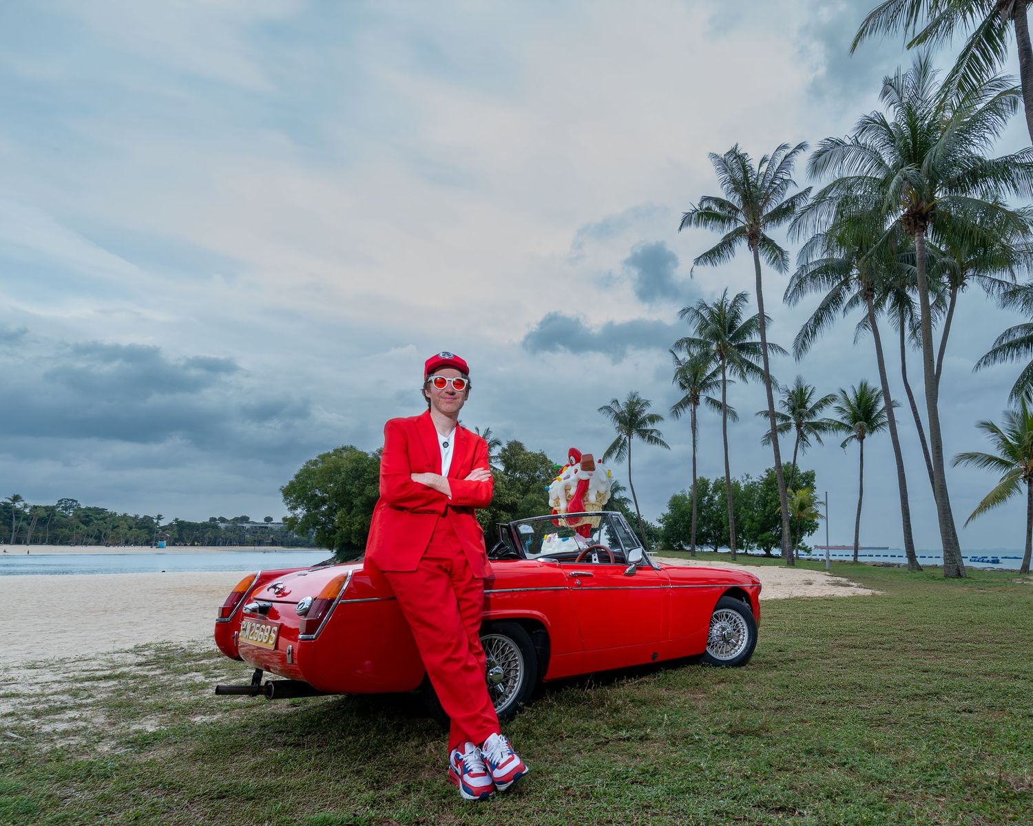 Person in red suit leaning on a classic red convertible car on a tropical beach with palm trees.