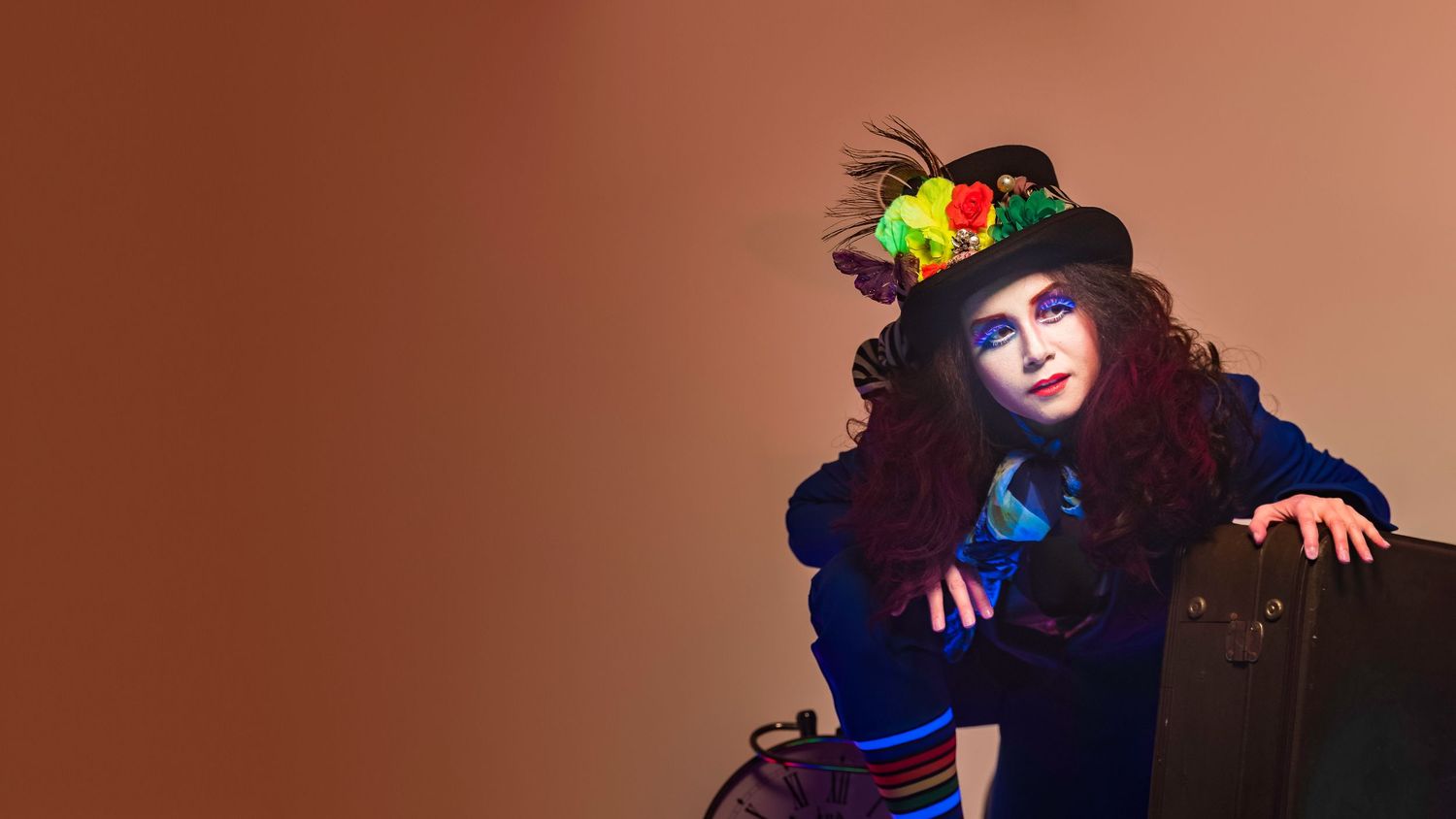 A whimsical woman in colorful Mad Hatter costume with floral hat and curly hair poses against brown background.
