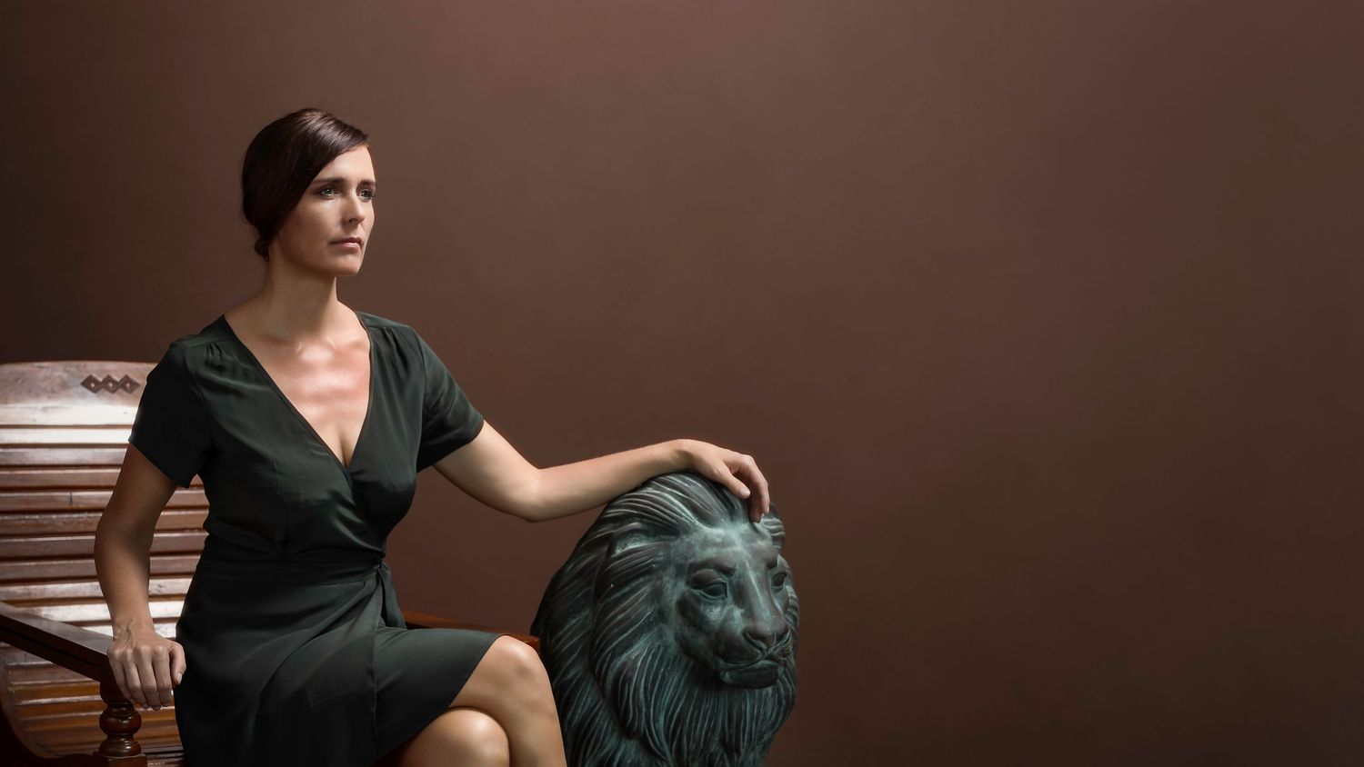 Elegant woman in dark green dress seated beside ornate lion sculpture against brown background.