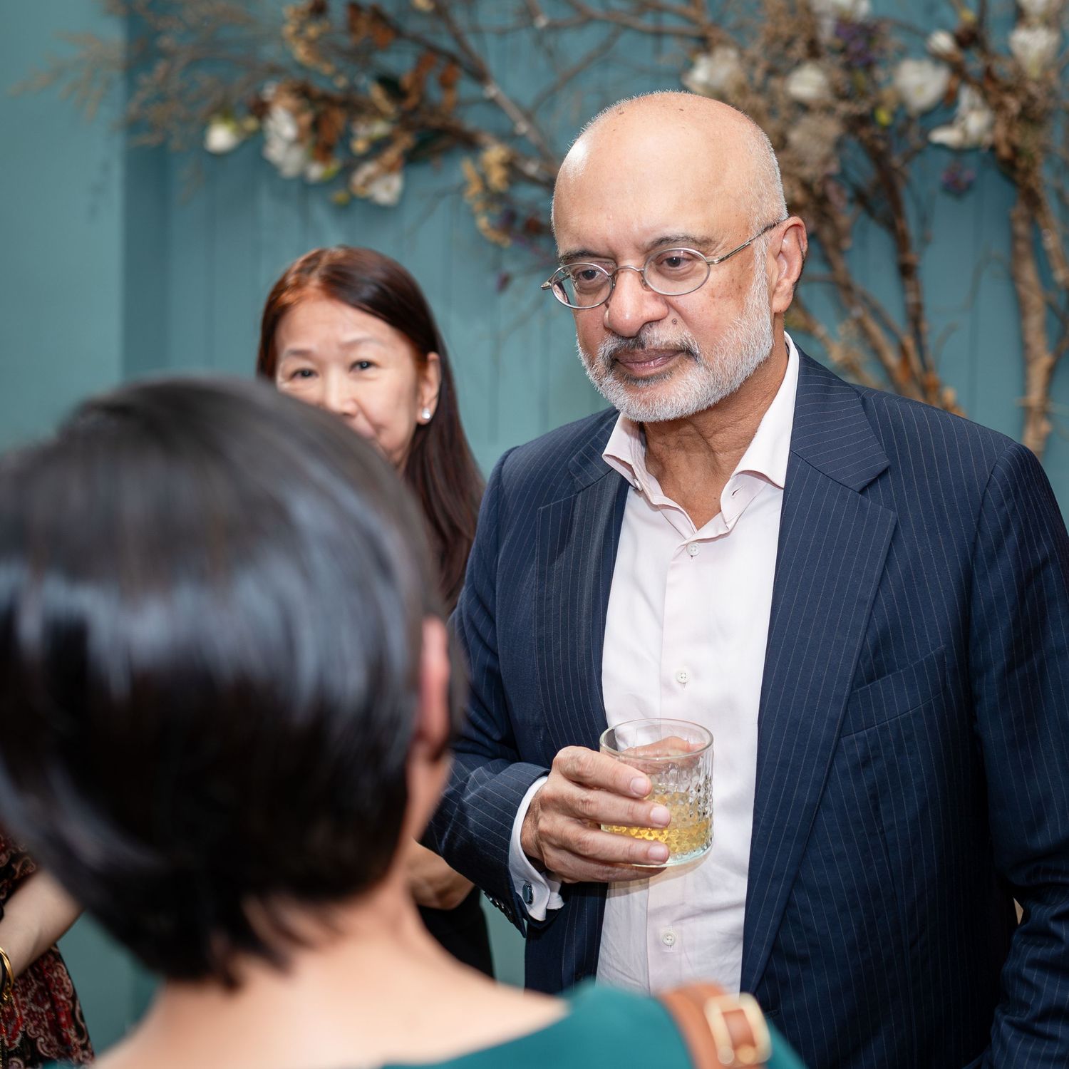 A bald man in a navy blazer holds a drink at an elegant social gathering with guests in the background.