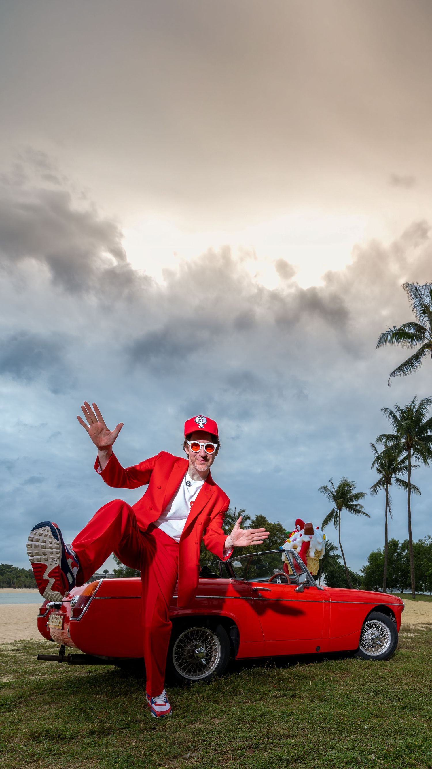 Man in all-red suit and cap posing on vintage red convertible car near tropical beach.