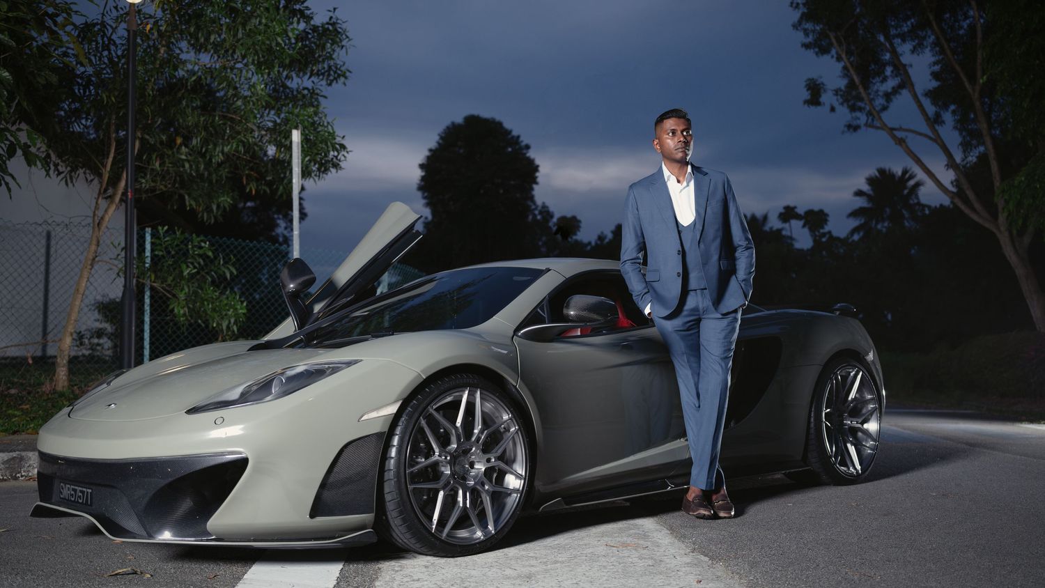 Stylish man in blue suit posing beside a sleek gray McLaren sports car at dusk.
