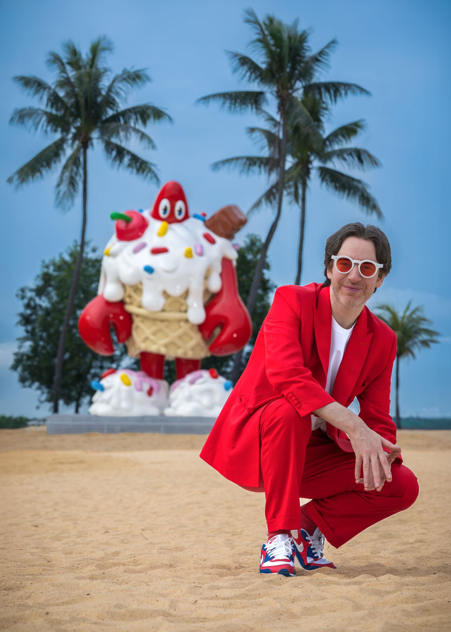 Person in red suit crouching on beach before giant ice cream cone sculpture with palm trees.